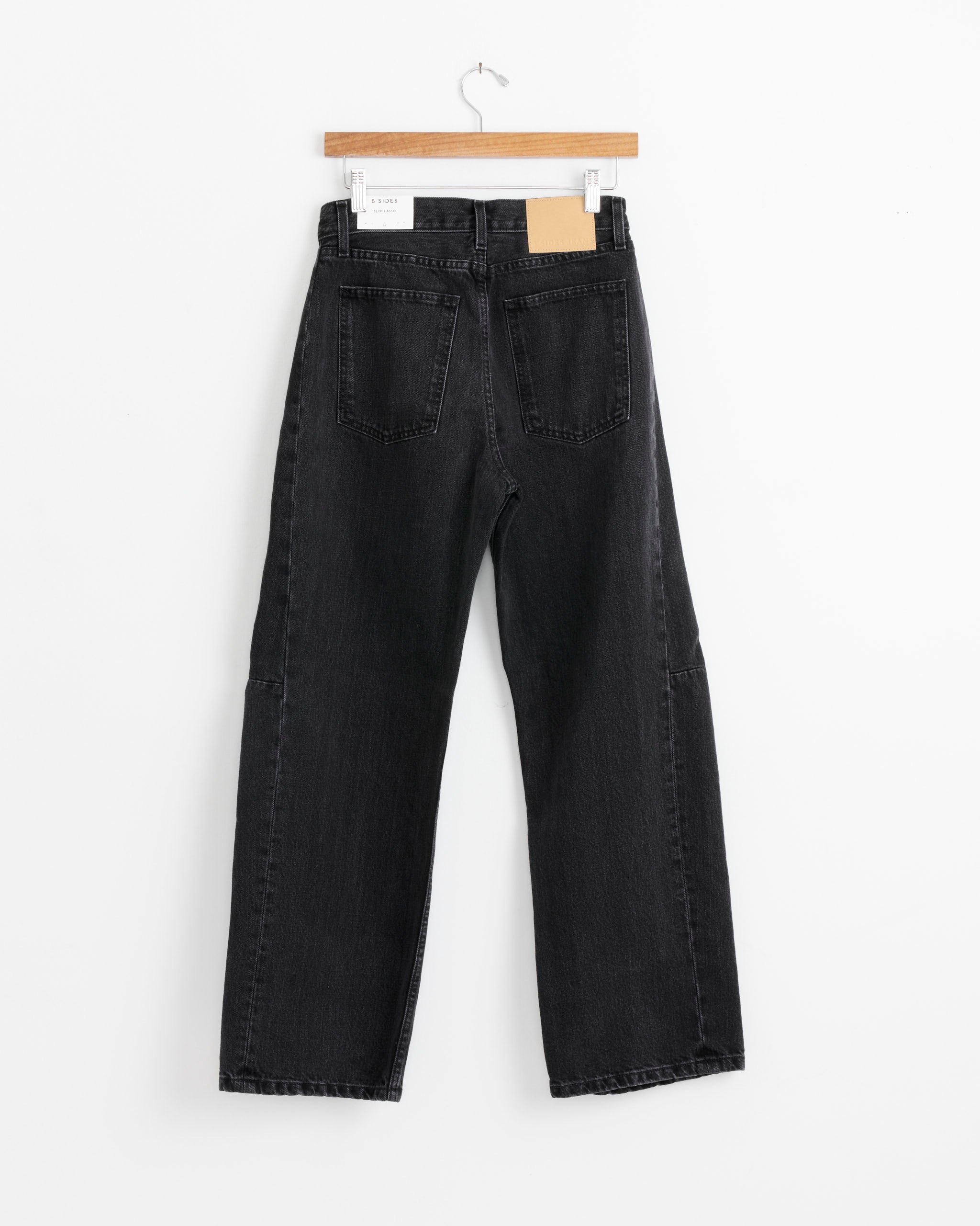 The B Sides Slim Lasso in Stil Black, a pair of high-waisted black denim jeans with straight legs and two back pockets, hangs on a wooden hanger against a plain white background.