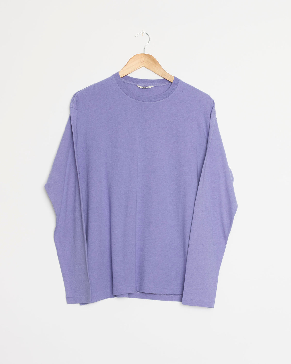 The Auralee Brushed Cotton Jersey Tee in Light Purple, crafted from soft brushed cotton jersey, hangs on a wooden hanger against a white background.