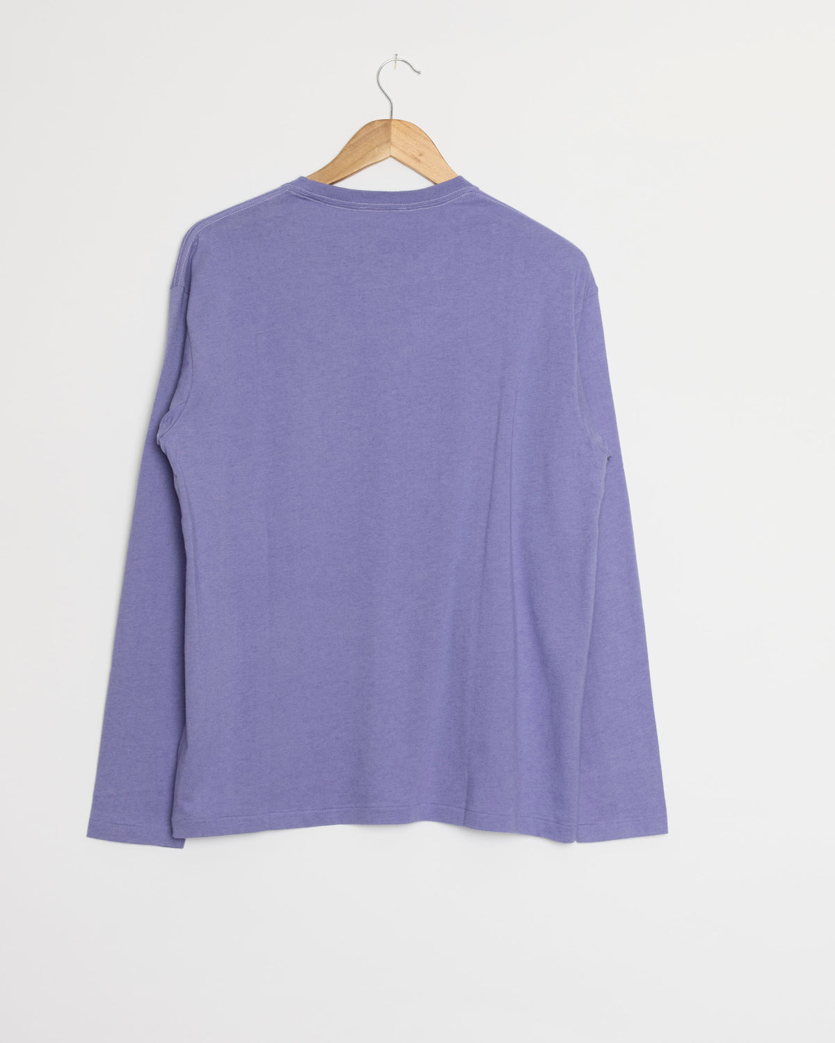 A Brushed Cotton Jersey Tee in Light Purple by Auralee, featuring long sleeves, is displayed on a wooden hanger against a plain white wall, shown from the back.