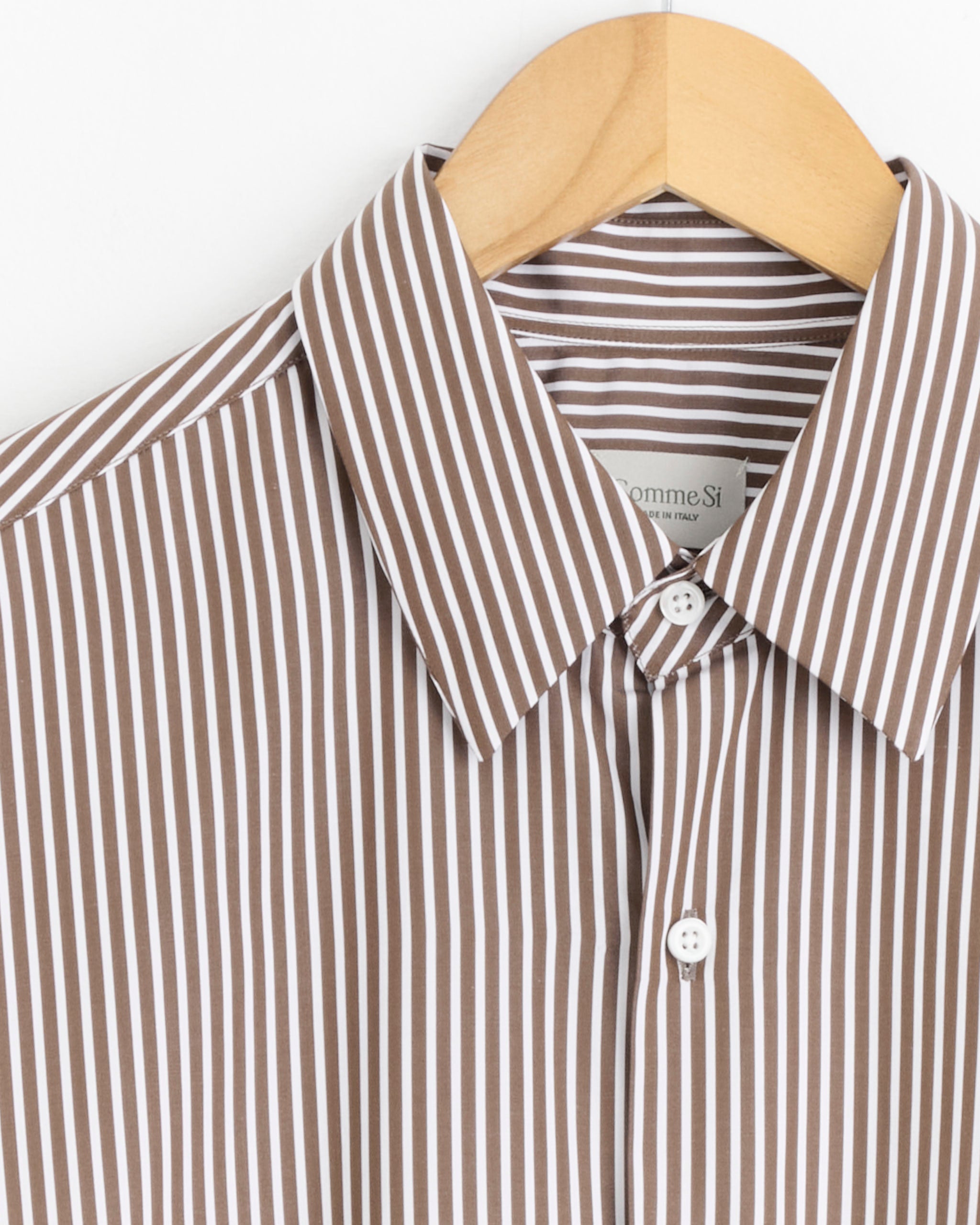A close-up of Comme Si’s La Shirt Classica in Cortado Stripe—a brown and white Italian cotton poplin button-up—hangs on a wooden hanger, collar, buttons, and part of the label visible.