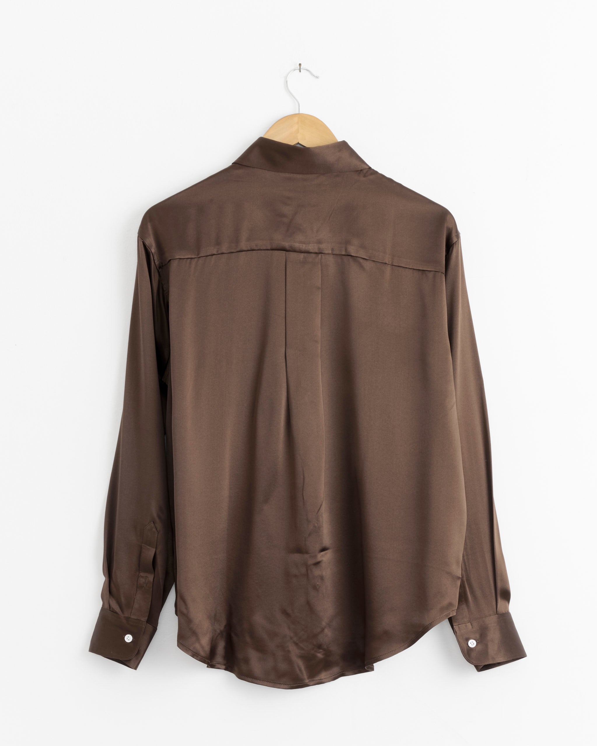 The Comme Si La Shirt Classica in Espresso, a unisex long-sleeve satin silk button-up, hangs from a wooden hanger against a white wall, shown from the back.
