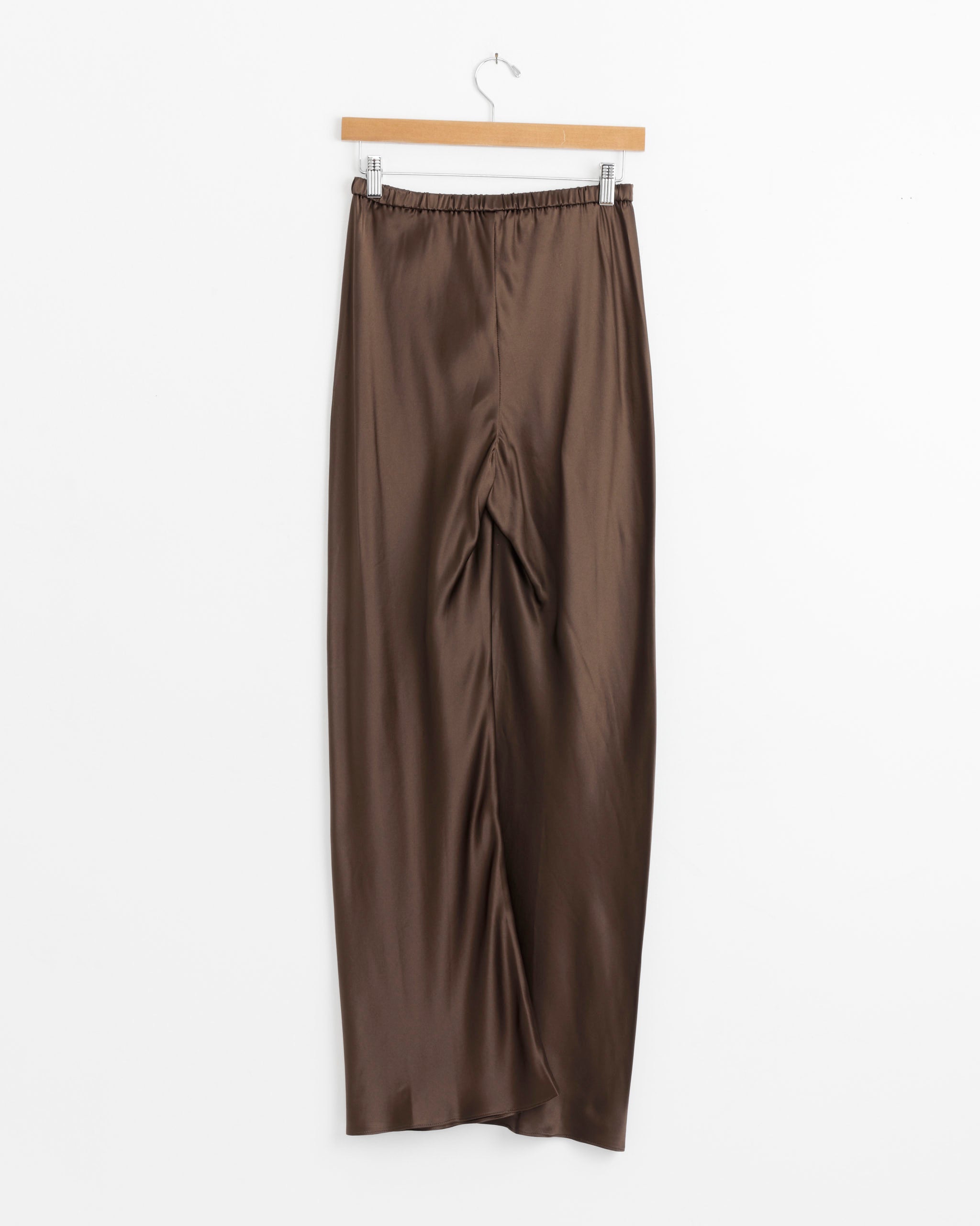 The Silk Bias Regular Pant in Espresso by Comme Si, featuring a wide-leg silhouette and elastic waistband, hangs on a wooden hanger against a plain white background.