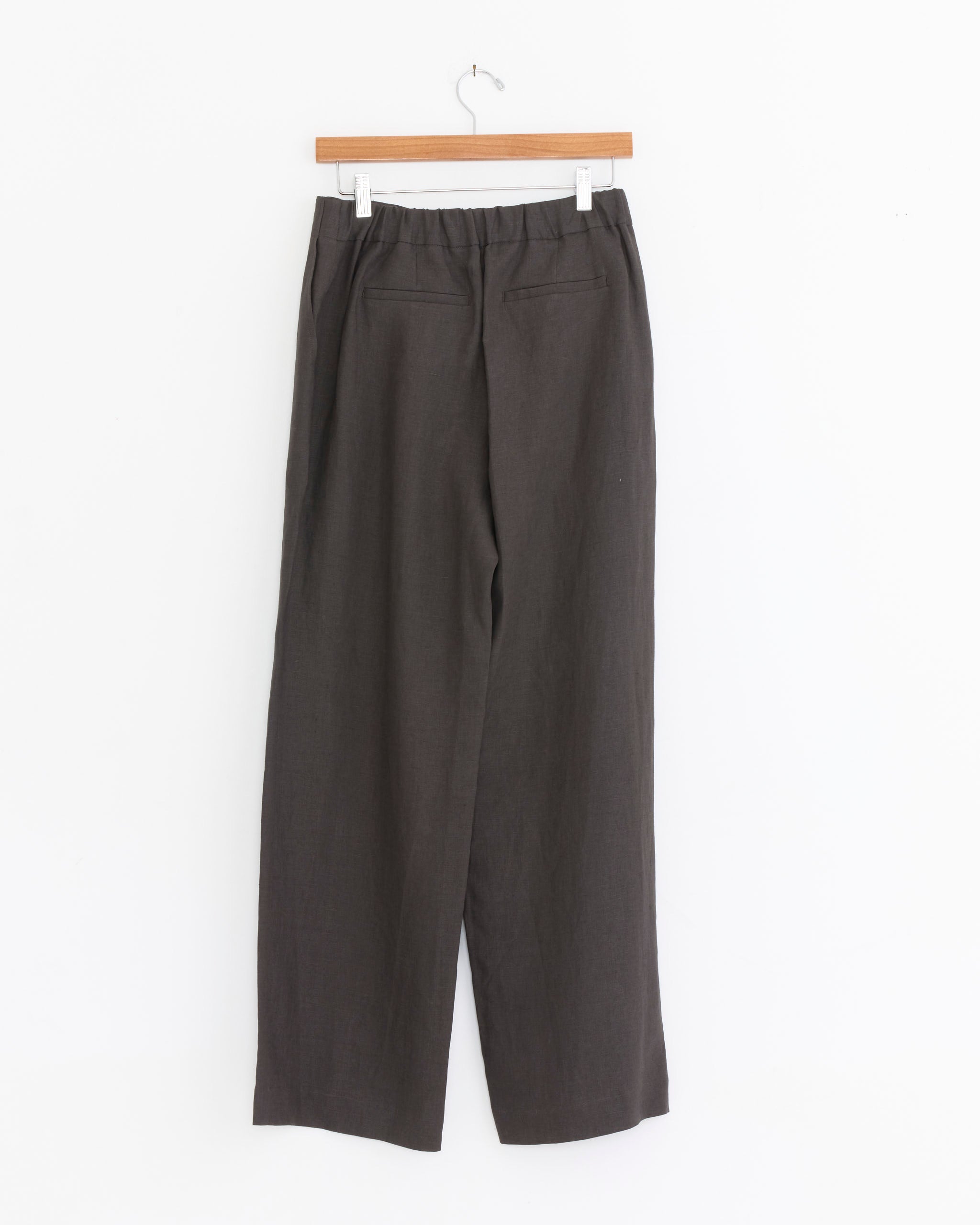 The Amomento Linen Tuck Pant in Khaki Grey features a straight fit, elastic waistband, and two back pockets, shown hanging on a wooden hanger against a white background.