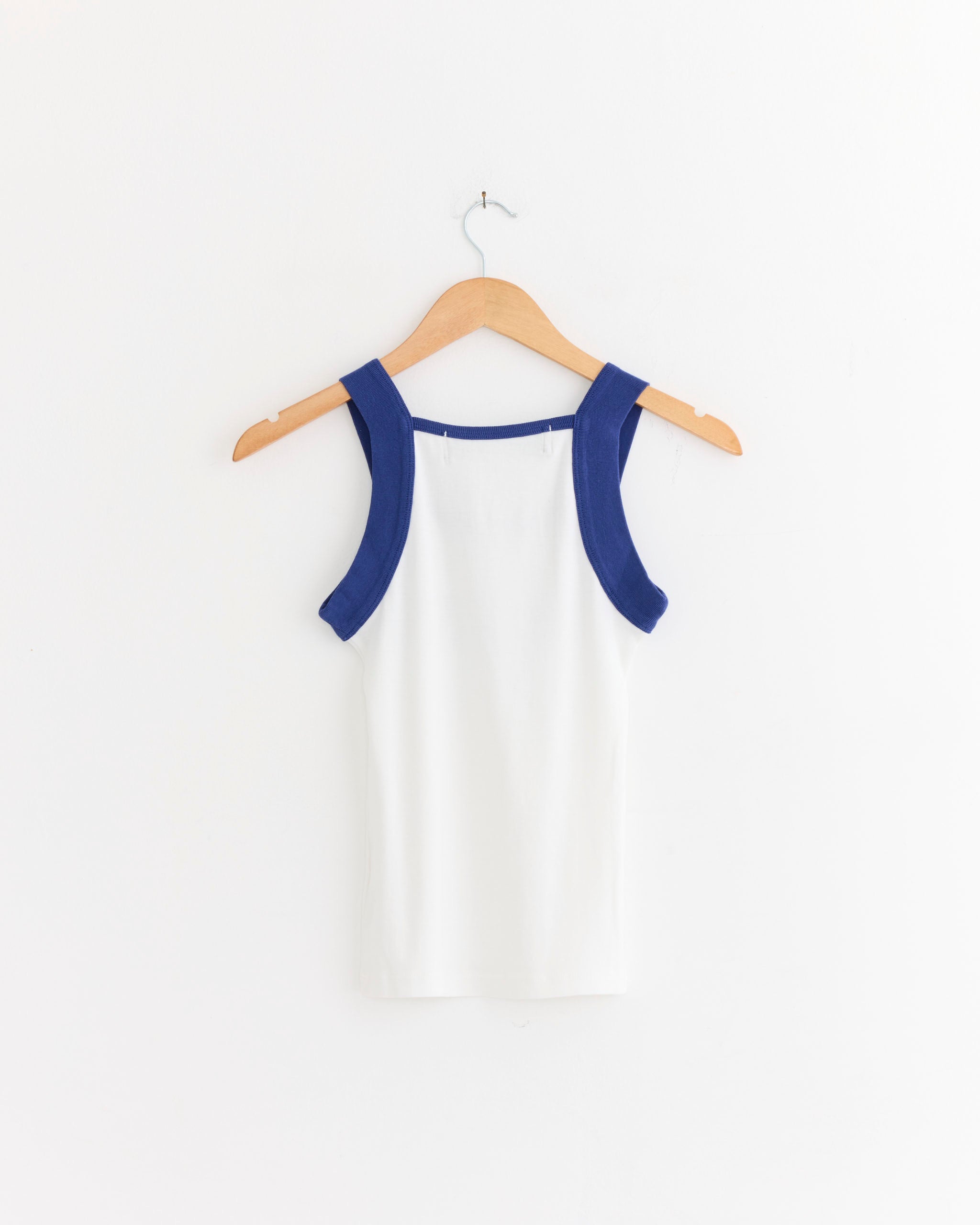 The Greece Tank Top in White/Blue by Made Some Sportswear, a sleeveless cotton ribbed top with wide blue straps, is displayed on a wooden hanger against a plain white wall.