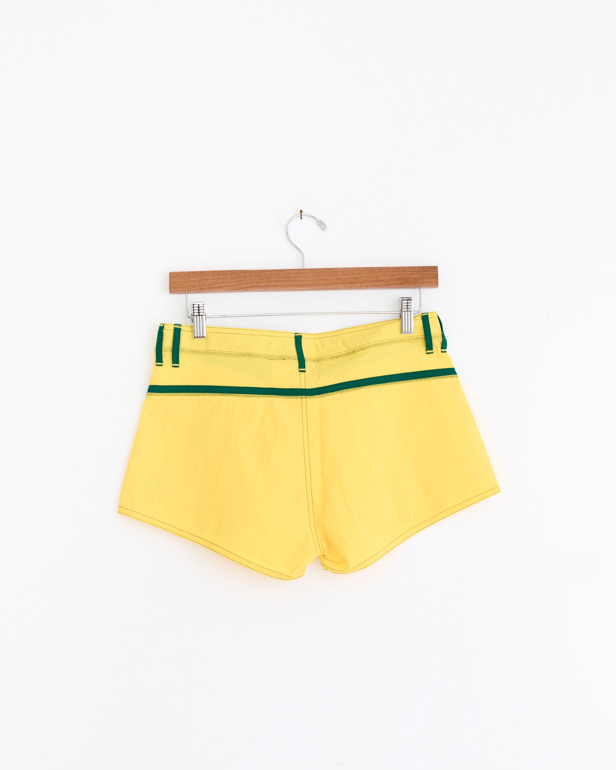 The Short Shorts in Yellow/Green by Made Some Sportswear feature lightweight waterproof nylon and green-striped waistband details, evoking classic 1970s surf style. Shown on a wooden hanger against a plain white background.