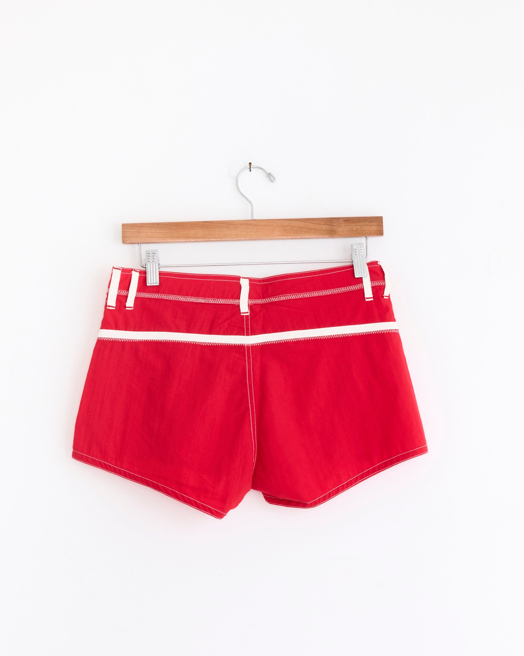 The Made Some Sportswear Short Shorts in Red/White, featuring classic '70s surf style and white trim, are displayed on a wooden hanger against a plain white wall.