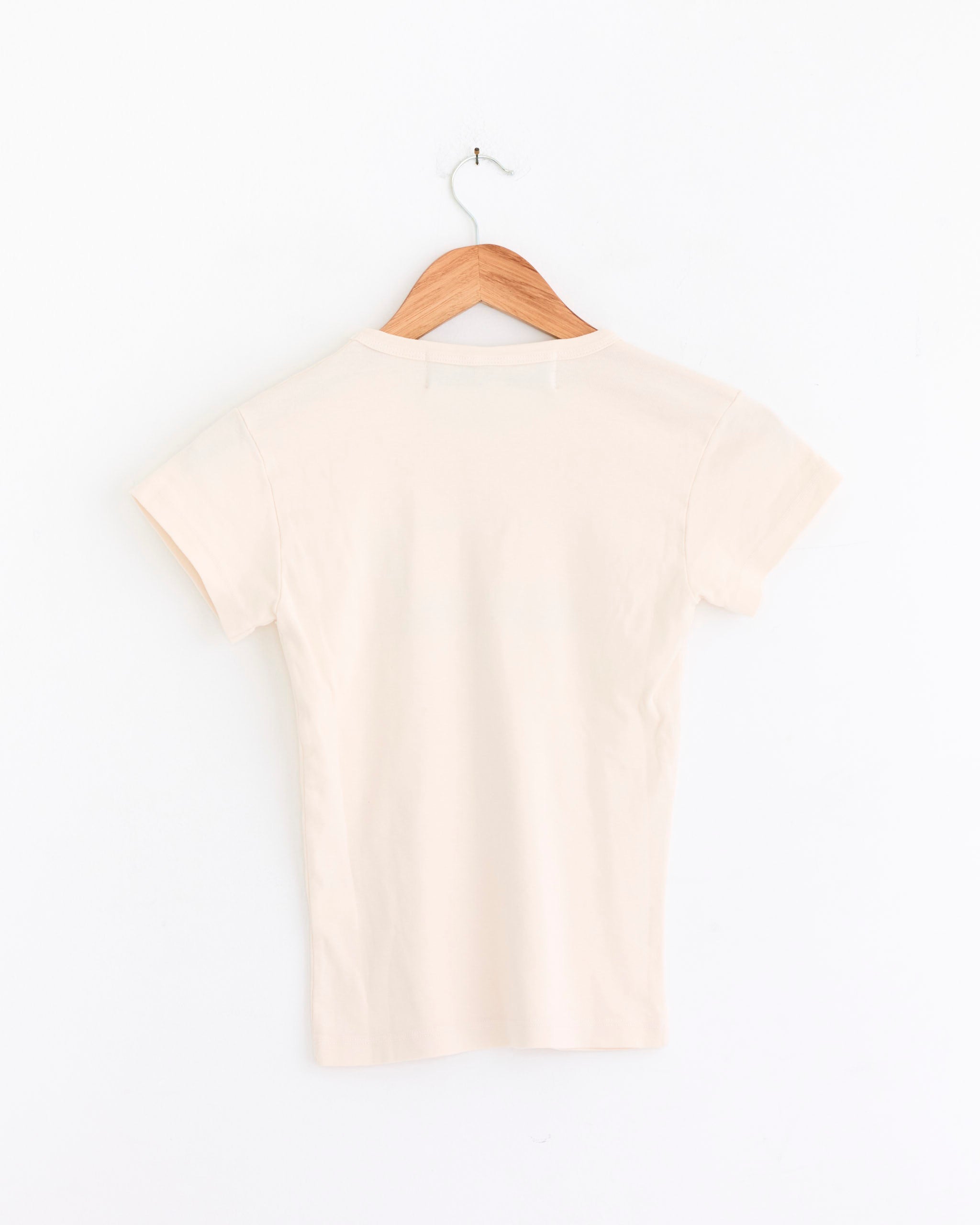 The Jamaica Tee in Multi by Made Some Sportswear, shown from the back, hangs on a wooden hanger against a white wall. This short-sleeved tee is crafted from USA-grown cotton in a light beige shade.
