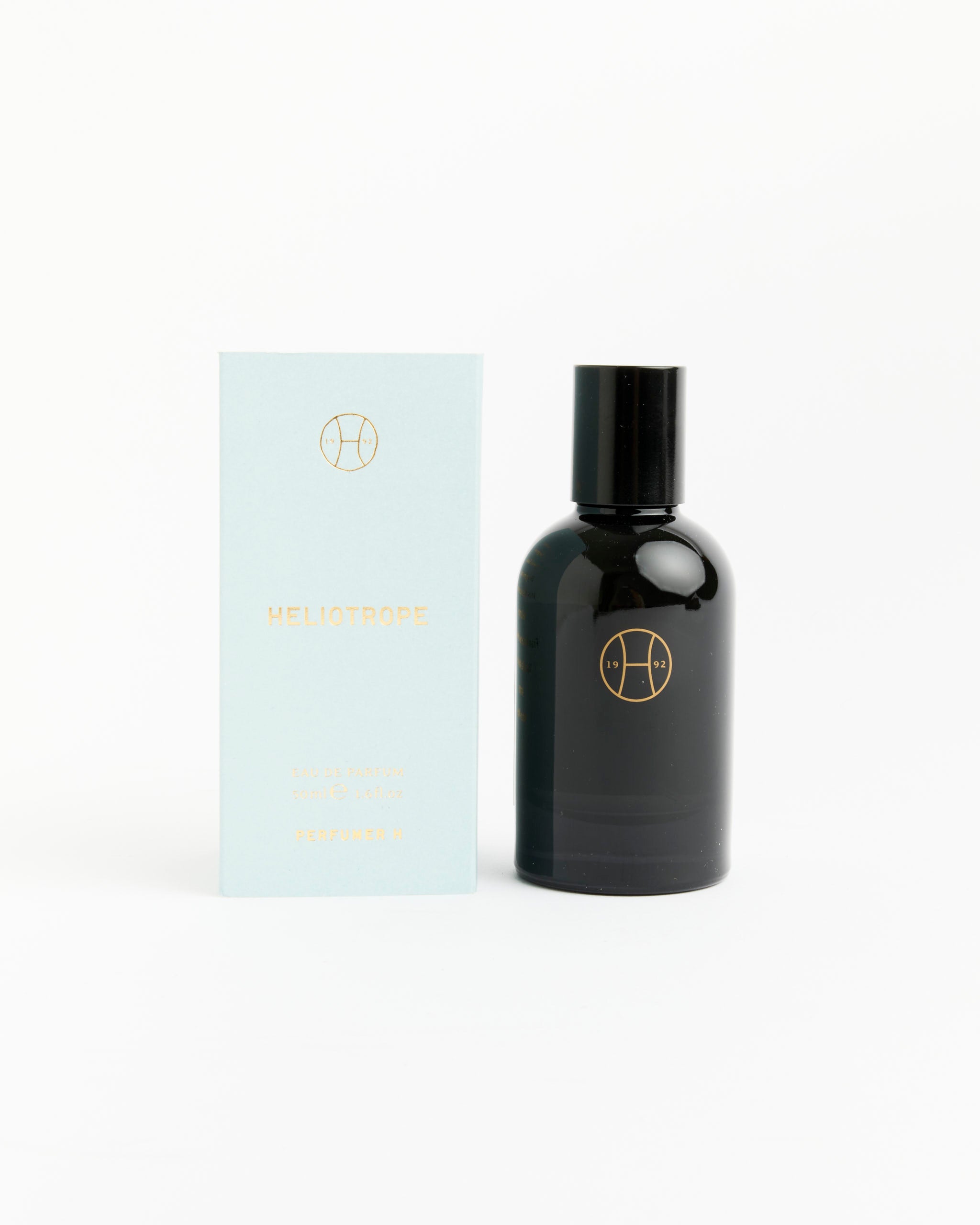 A light blue Perfumer H Heliotrope Perfume box stands beside a sleek black bottle with a gold logo on a white surface, both items forming a modern amber fragrance set.
