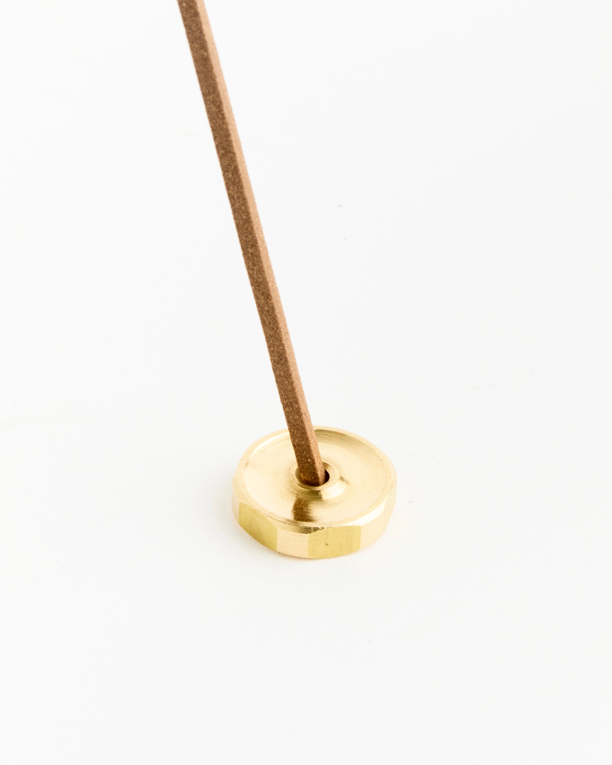 A brown hand-rolled incense stick from Perfumer H’s Gold Incense is held upright in a small, round, gold-colored metal holder on a white background.