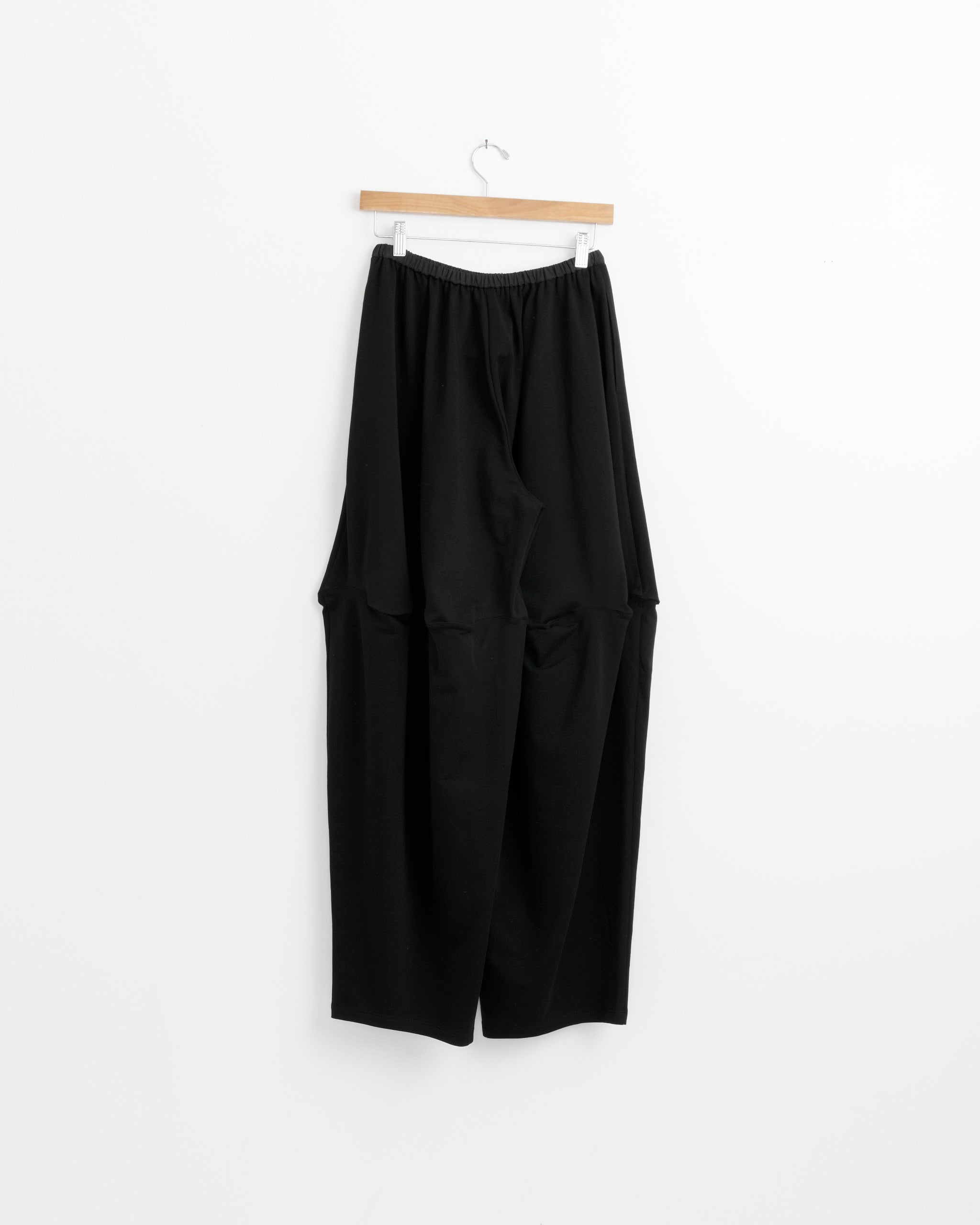 The J. Kim Bakeware Pant in Black, featuring a loose fit and elasticated waistband, hangs on a wooden hanger against a plain white background.