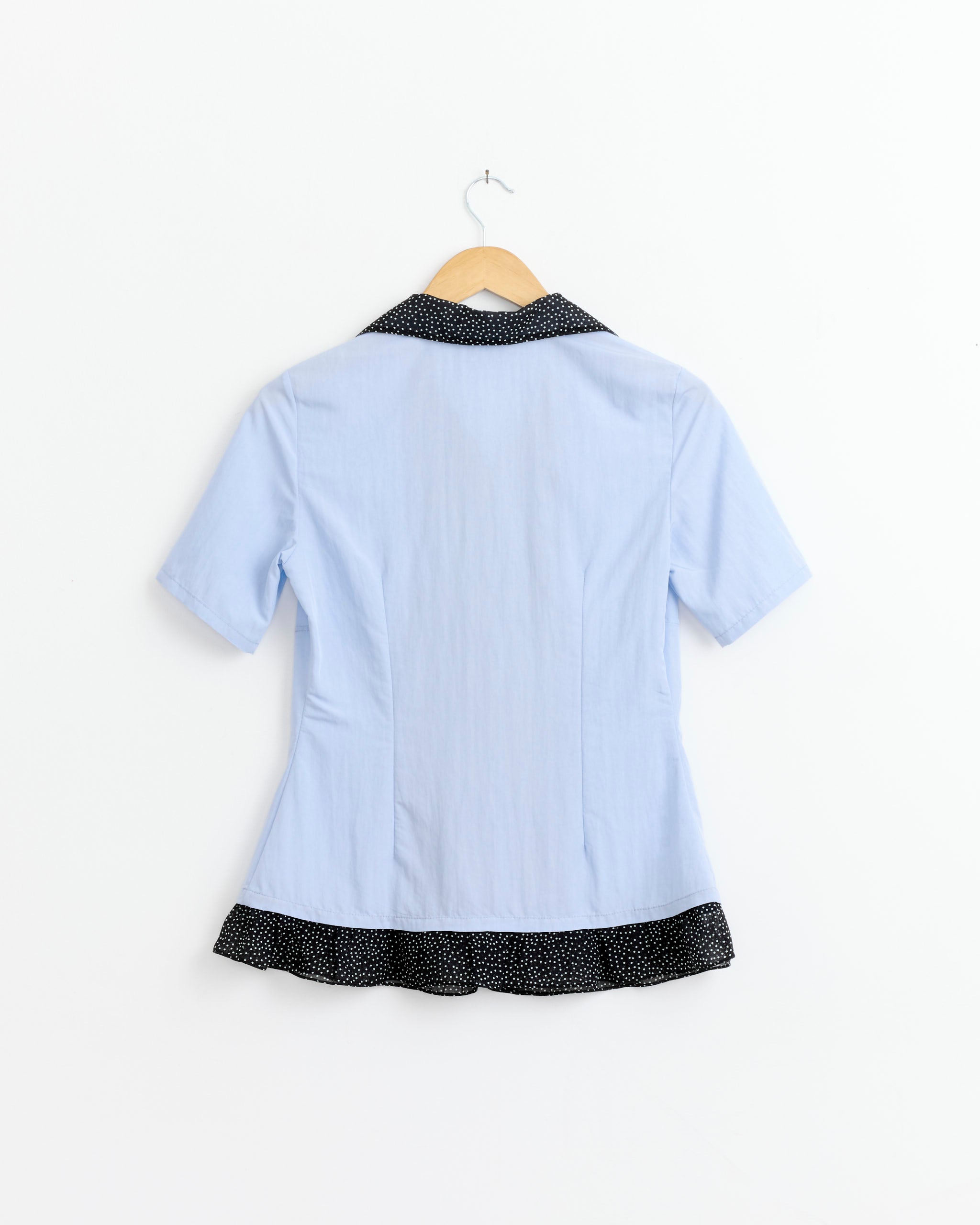 The Anechka Petal Shirt in Blue by J. Kim is a semi-fitted light blue shirt with a black collar and white polka-dot ruffle trim, shown on a wooden hanger against a white background.