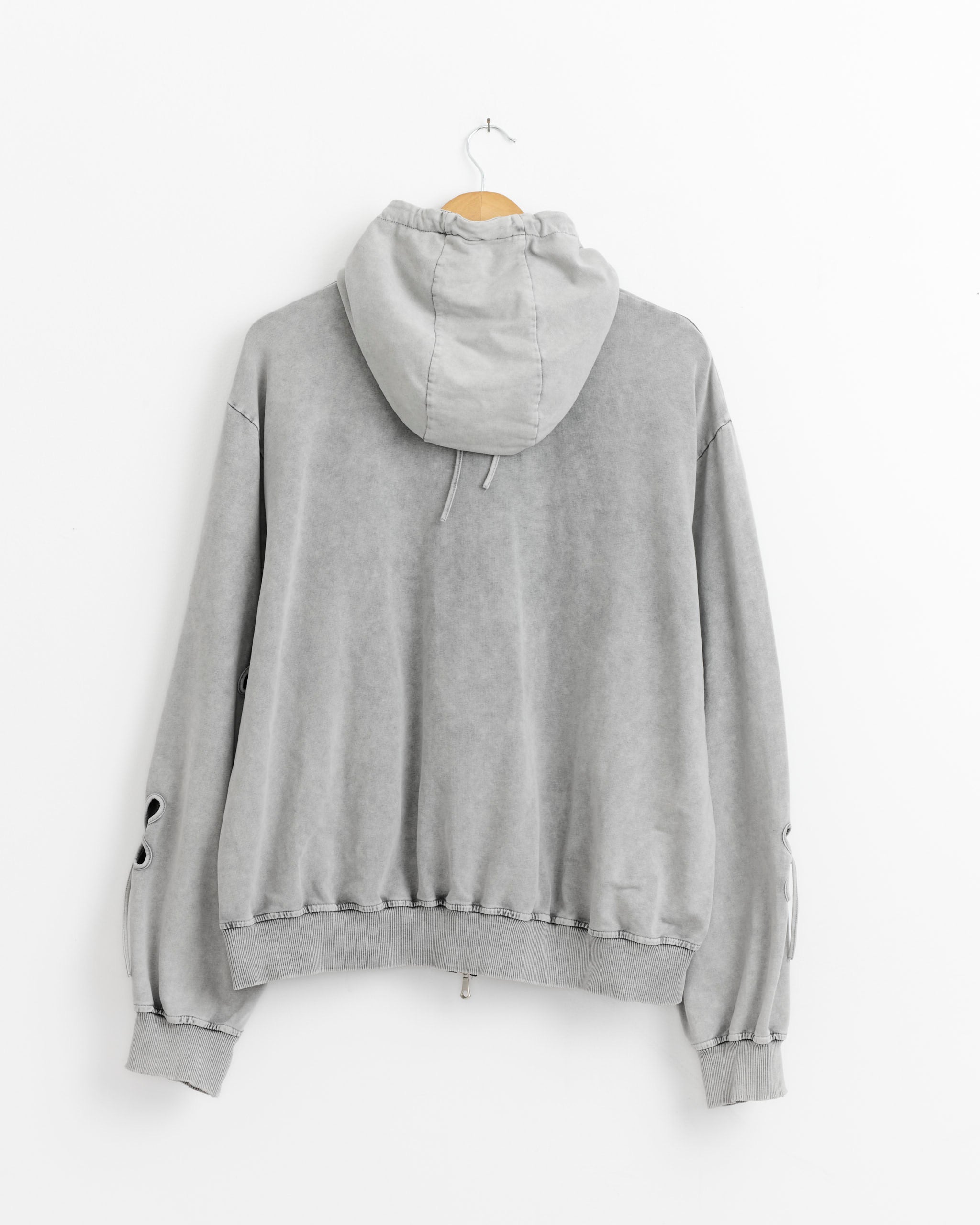 The Petal Hoodie in Ash Grey by J. Kim hangs on a wooden hanger against a white wall, displayed from the back with visible drawstrings and two small sleeve buttons.