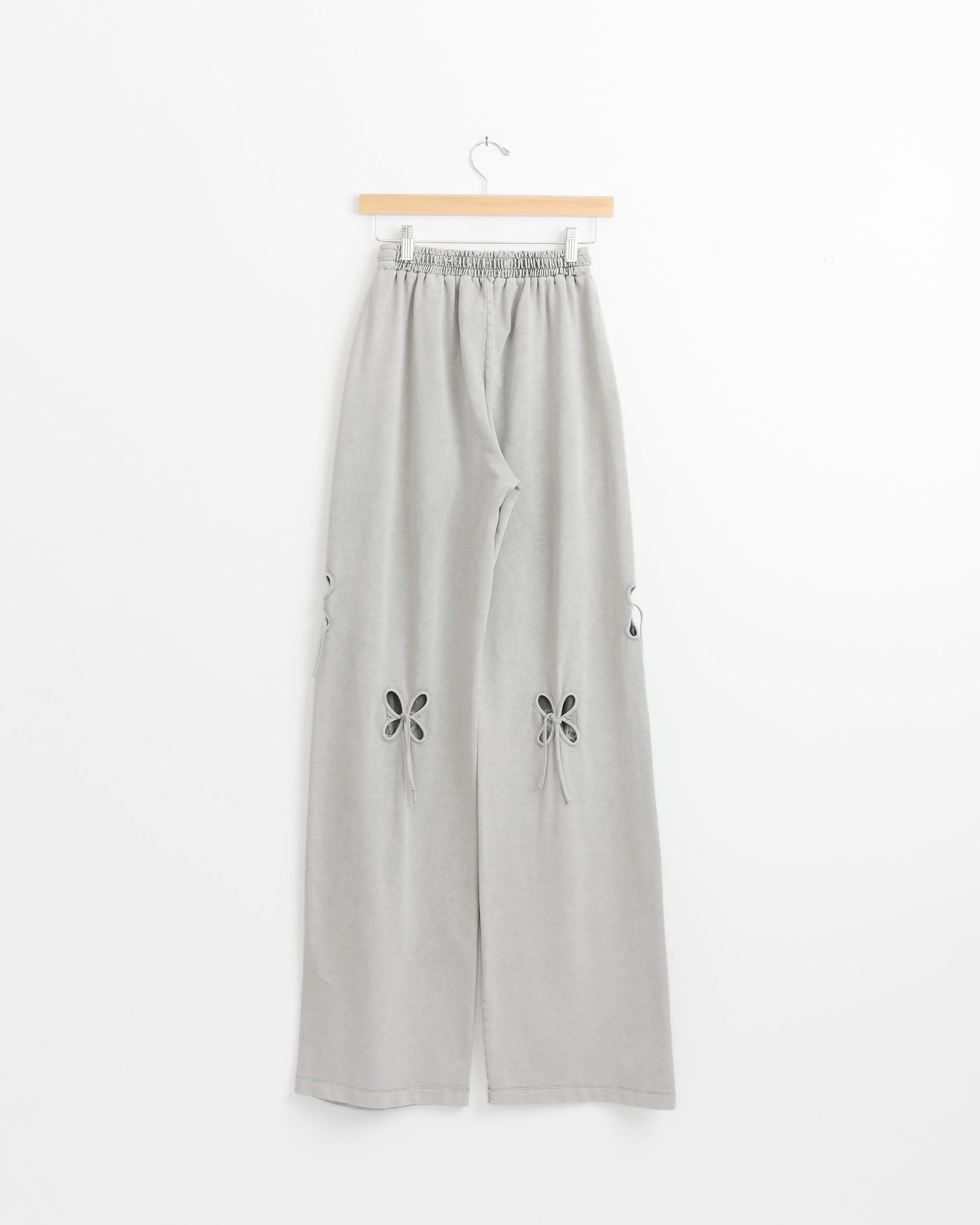 The Petal Pant in Ash Grey by J. Kim features an elastic waistband, cut-out flower details, and tied bows on each leg. Shown on a wooden hanger against a white background for a fresh, modern look.