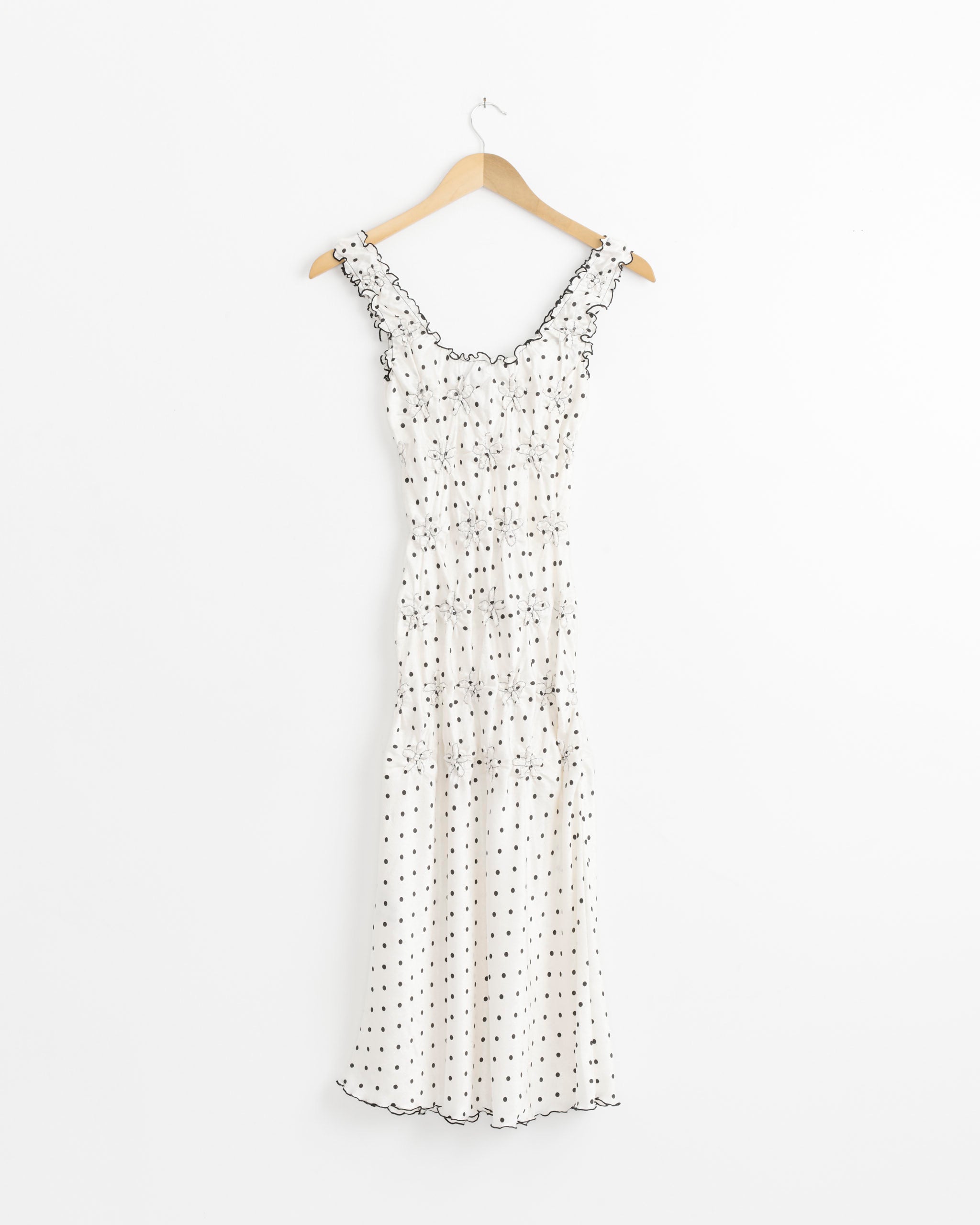 The Mariza Dress in Polka Dot by J. Kim is a sleeveless white midi dress with black polka dots, ruffled straps, and a fitted bodice, displayed on a wooden hanger against a plain white background.