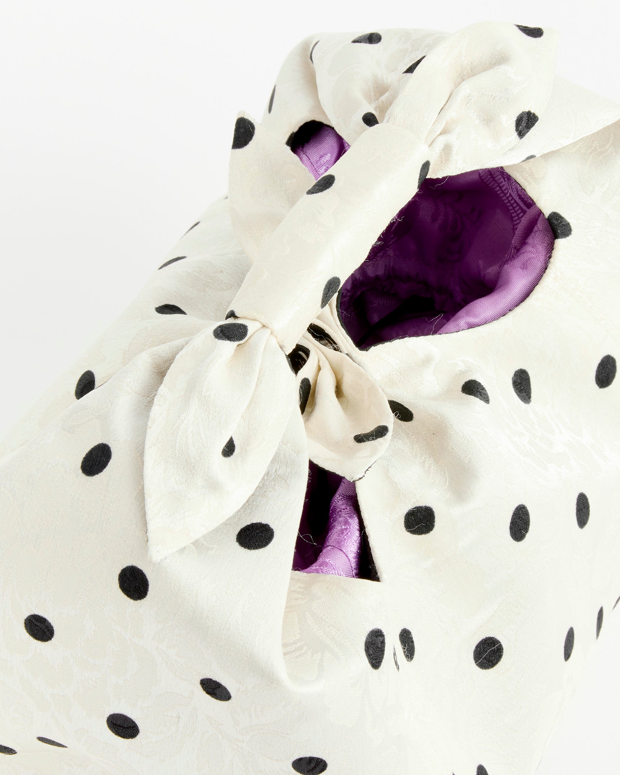 The Tugun Bag in Polka Dot by J. Kim features a white polka dot design, tied at the top in a bow, with a peek of purple lining—a modern twist on the classic Uzbek bundle.