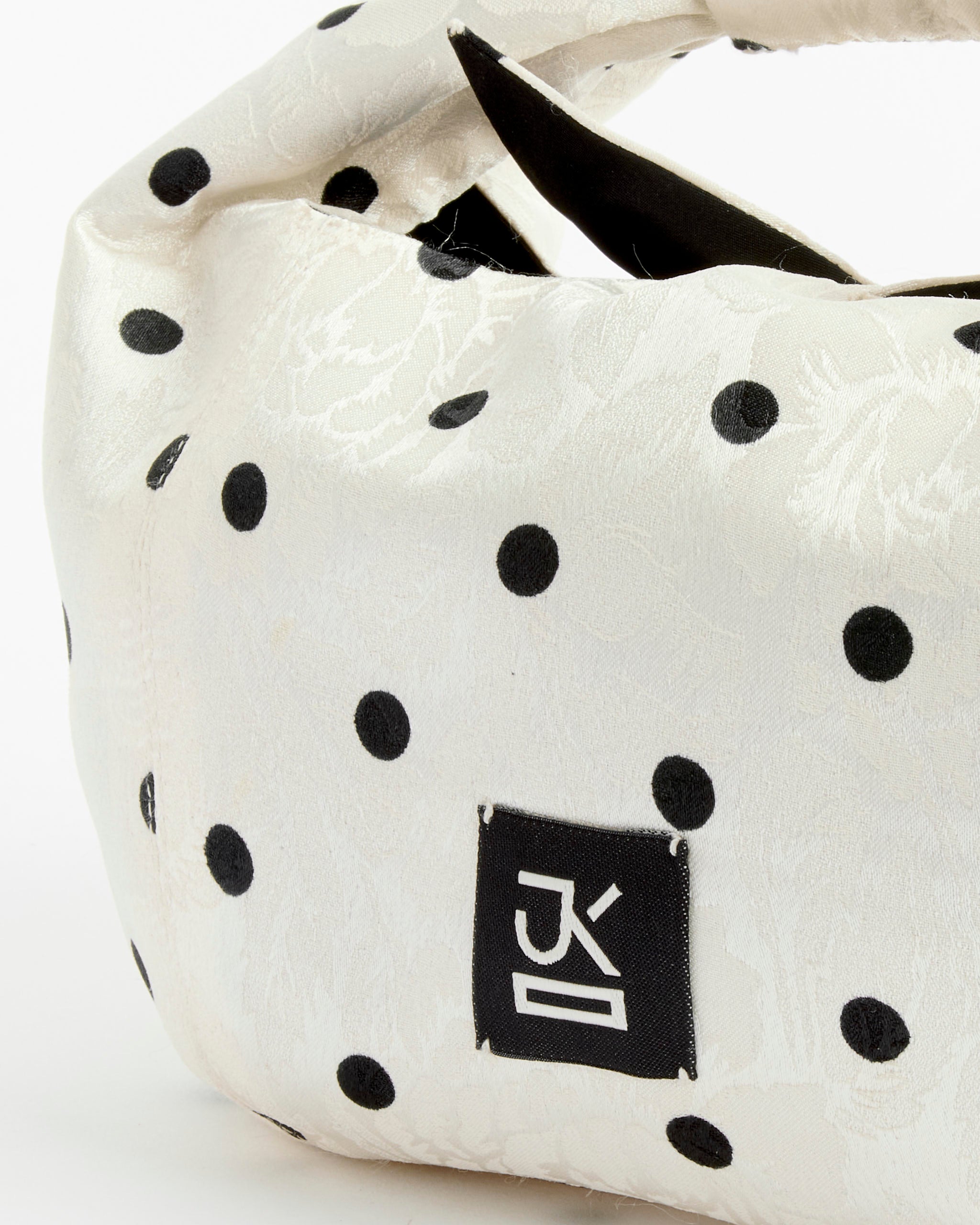 A close-up of the Tugun Bag in Polka Dot by J. Kim, an Uzbek bundle crafted from white fabric with black polka dots and a floral texture, featuring a black rectangular label with white stylized text near the bottom.