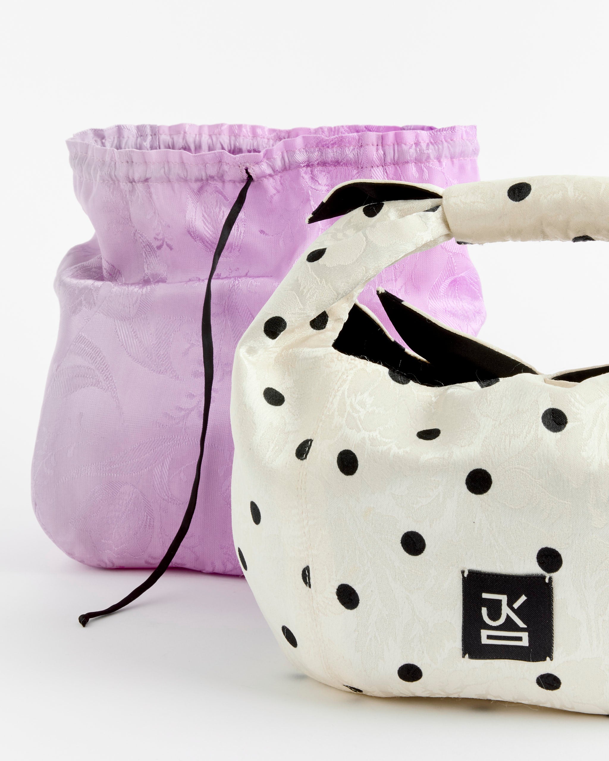 A J. Kim Tugun Bag in Polka Dot with a knotted handle is displayed in front of a light purple drawstring pouch on a white background.