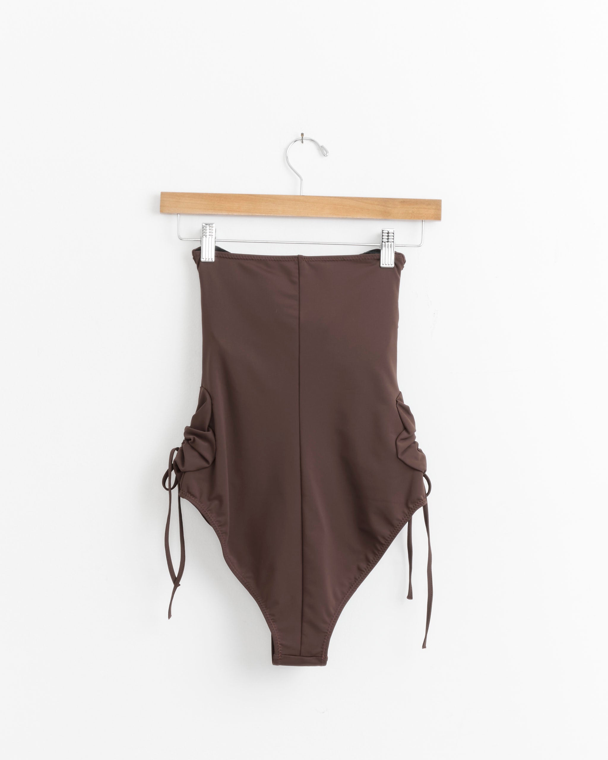 The Flower One Piece in Cinnamon by J. Kim is a brown, high-waisted, strapless swimsuit with ruched sides and adjustable drawstrings, made from recycled polyamide, shown on a wooden hanger against a plain white background.
