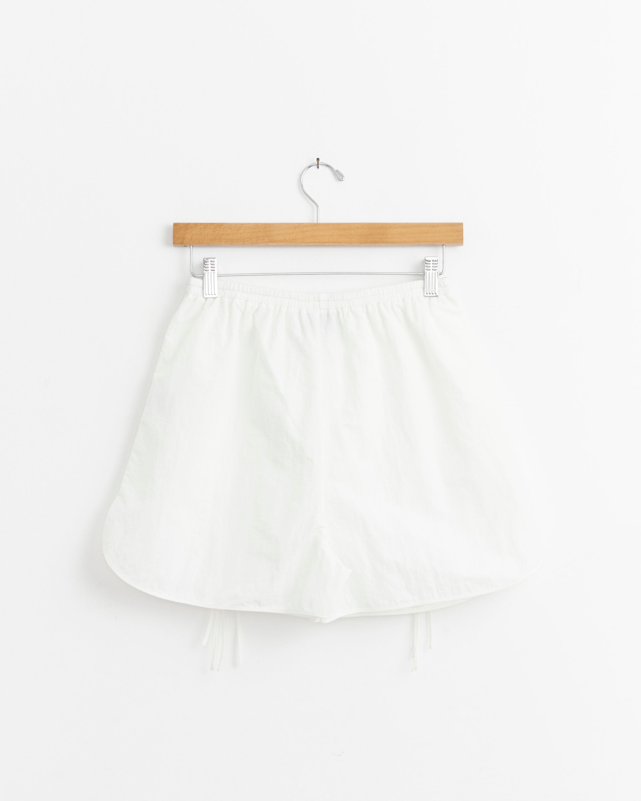 The Petal Short in White by J. Kim, featuring a relaxed fit and elastic waistband in lightweight nylon taffeta, hangs on a wooden hanger against a plain white wall.