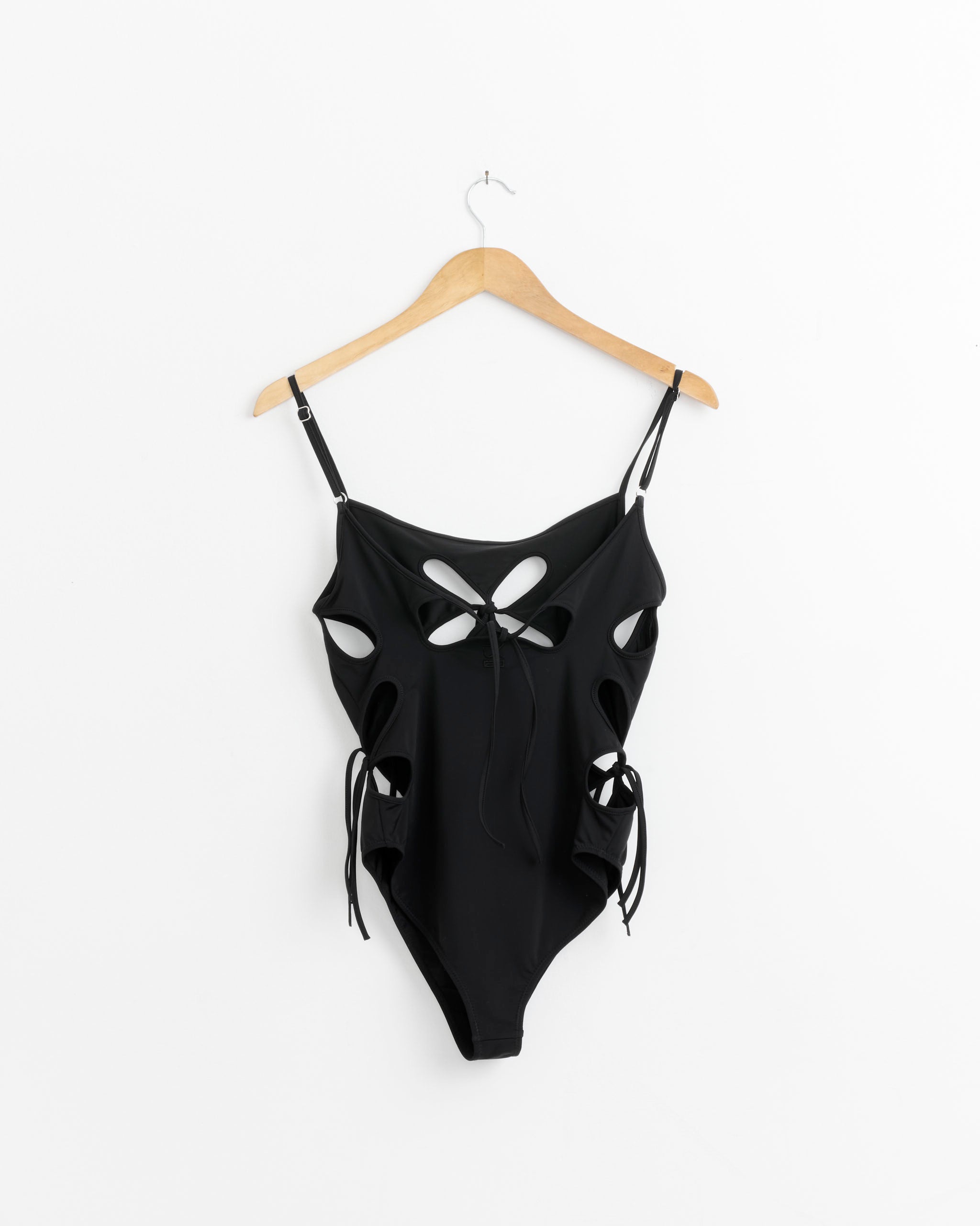 The Chilla Elongated Petal One Piece in Black by J. Kim features recycled fabric, multiple cutouts, side ties, and UV protection—shown hanging on a wooden hanger against a white background for a stylish look.