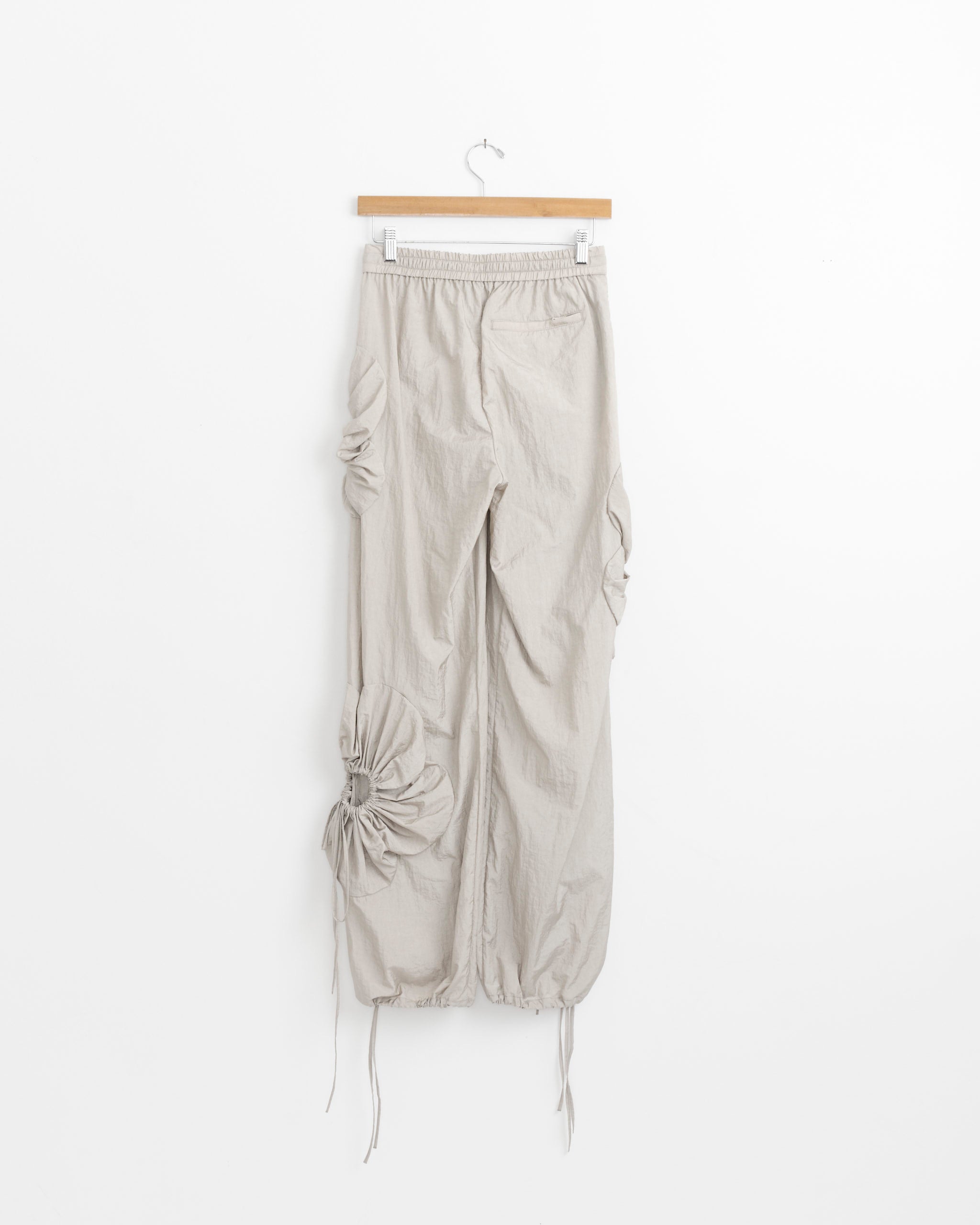 The Flower Pant in Beige by J. Kim hangs on a wooden hanger, showcasing ruched and gathered side and lower leg details, set against a plain white background.