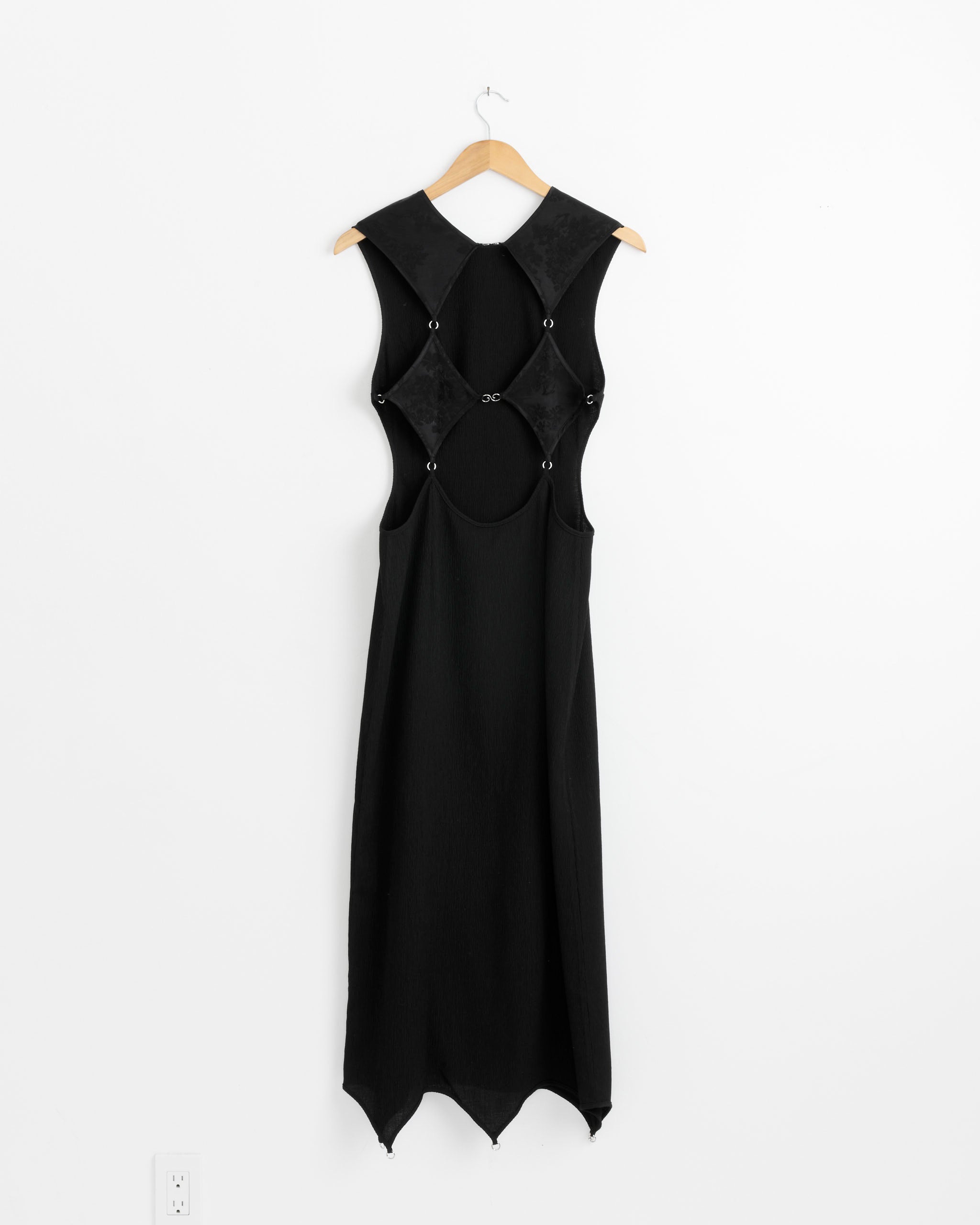The Lattice Dress in Black by J. Kim hangs on a wooden hanger against a white wall, showcasing its sleeveless design, diamond-shaped side cutouts on the bodice, button details, and an asymmetrical hemline.