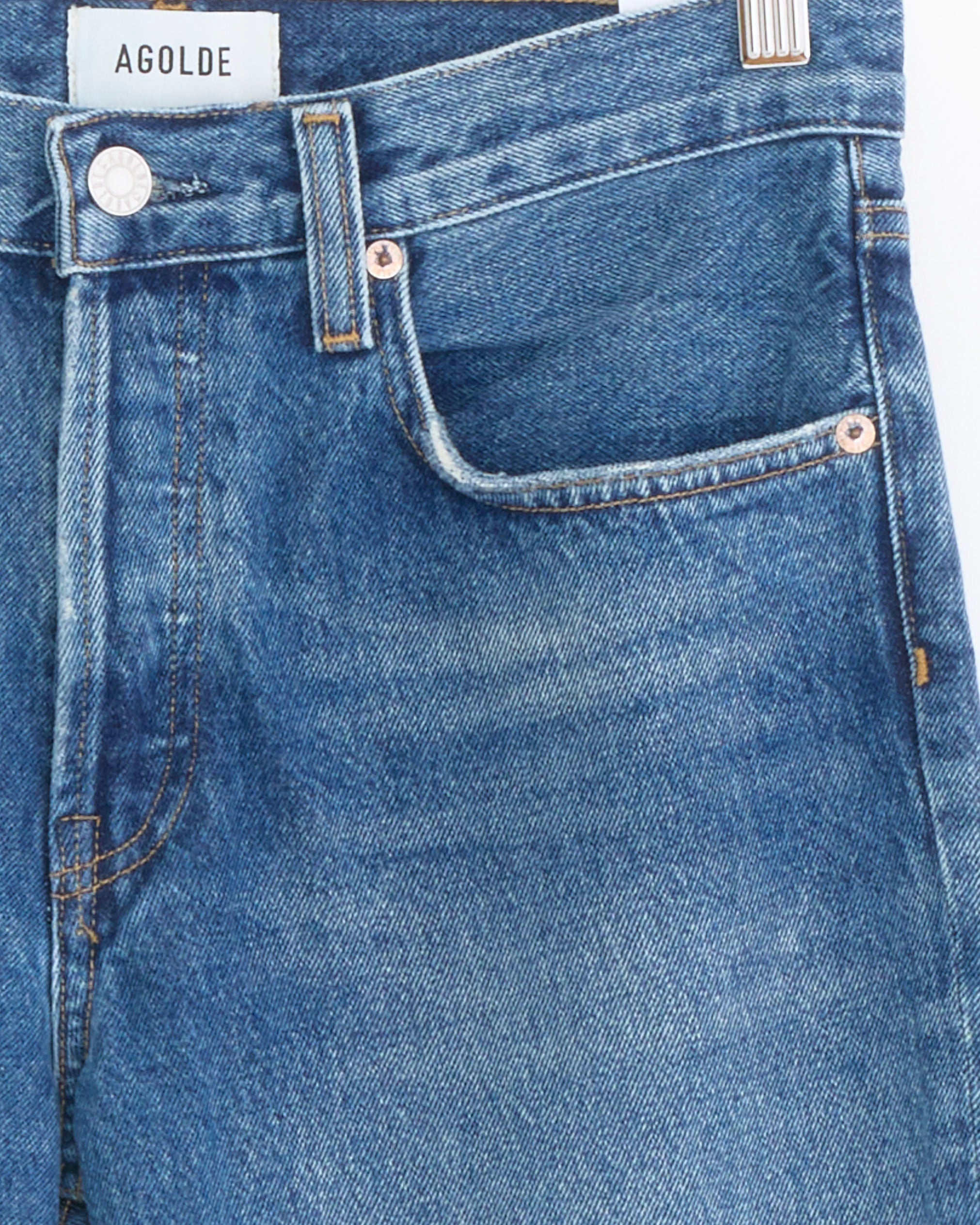 Close-up of Romney Jean in Bounty by AGOLDE, mid-rise blue denim with visible stitching, metal button, and front pocket. The white AGOLDE label is sewn on the waistband. Made from regenerative cotton denim and shown against a white background.