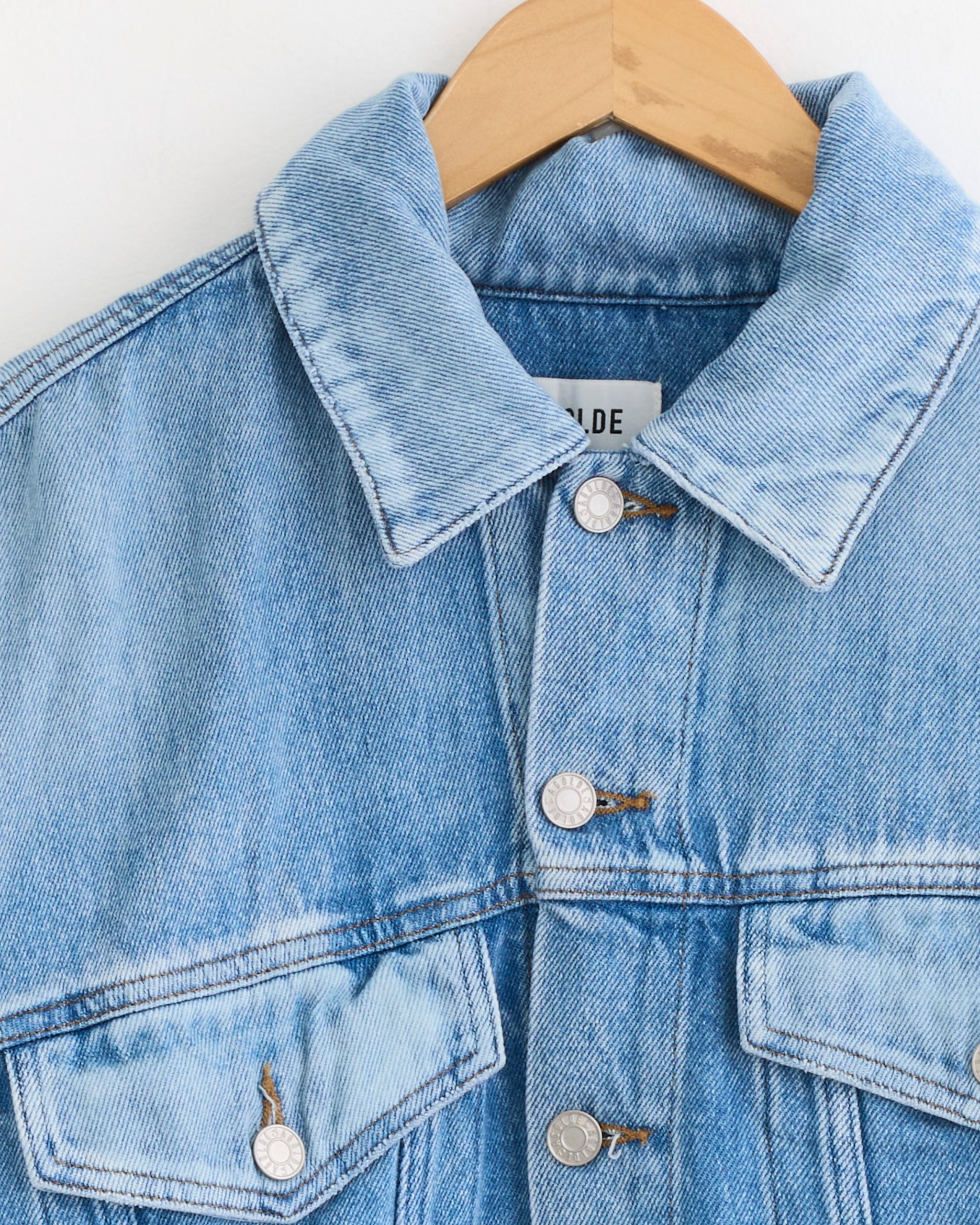 The Palmer Jacket in Script by AGOLDE—a cropped light blue denim jacket made from regenerative cotton with a collar, silver buttons, and two chest pockets—hangs on a wooden hanger against a plain white background.
