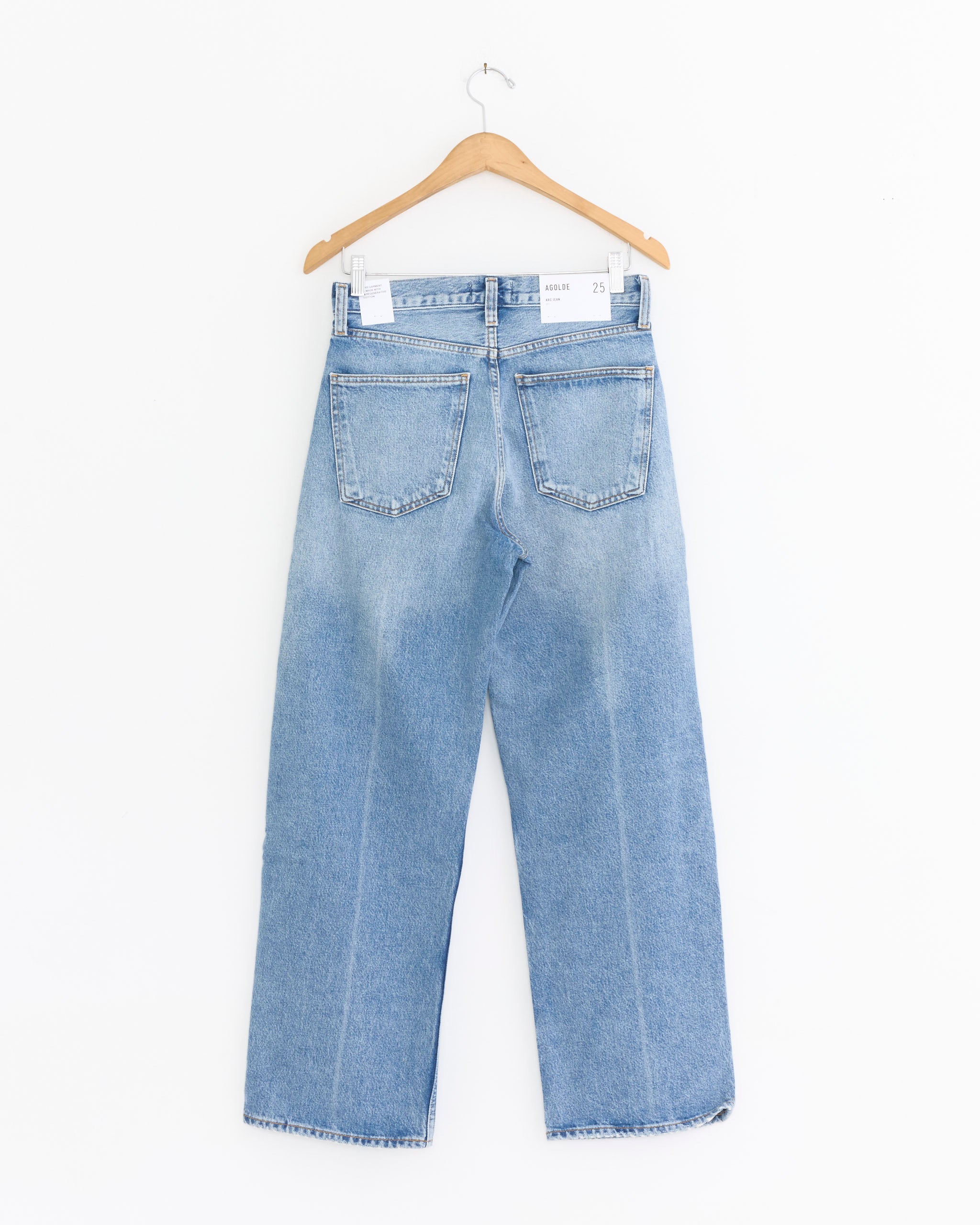 The AGOLDE Arc Jean 29" in Brink, a high-waisted light blue wide-leg style with two back pockets, is displayed on a wooden hanger against a plain white background.