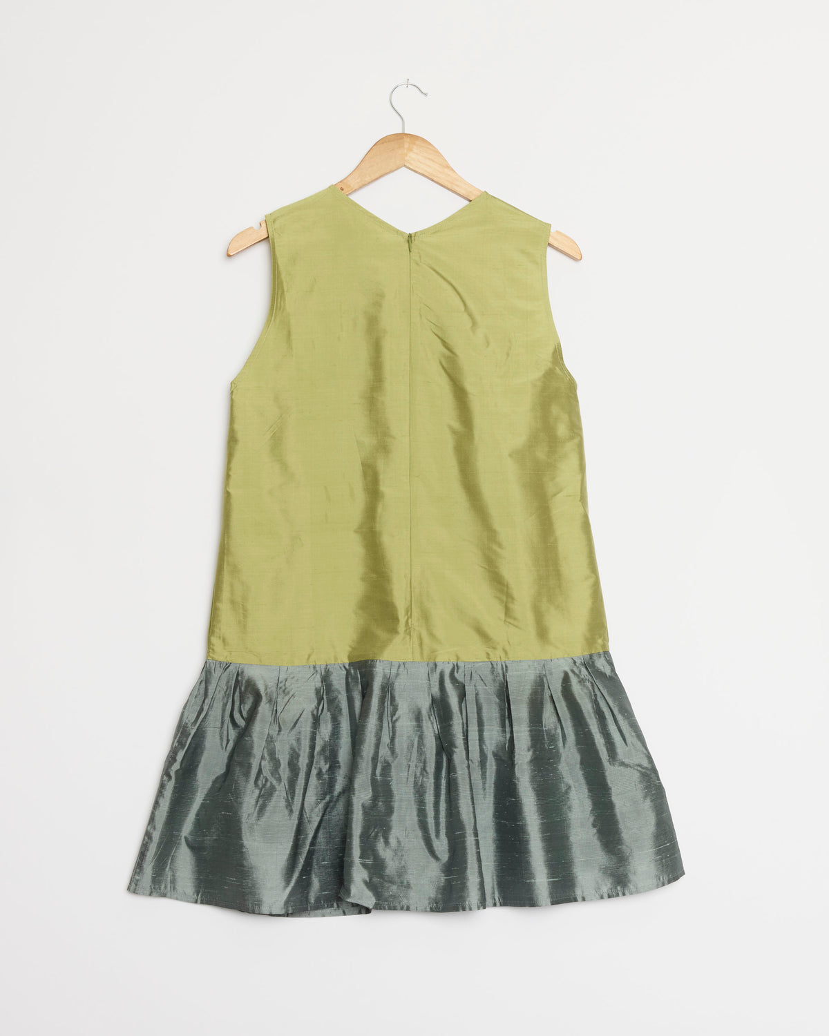 The Florence Dress in Multi by Chelsea Mak hangs on a wooden hanger, showcasing its sleeveless design with a light green bodice and a shiny, ruffled metallic gray skirt against a plain white background. For questions about fit or sizing, feel free to reach out via our online chat.