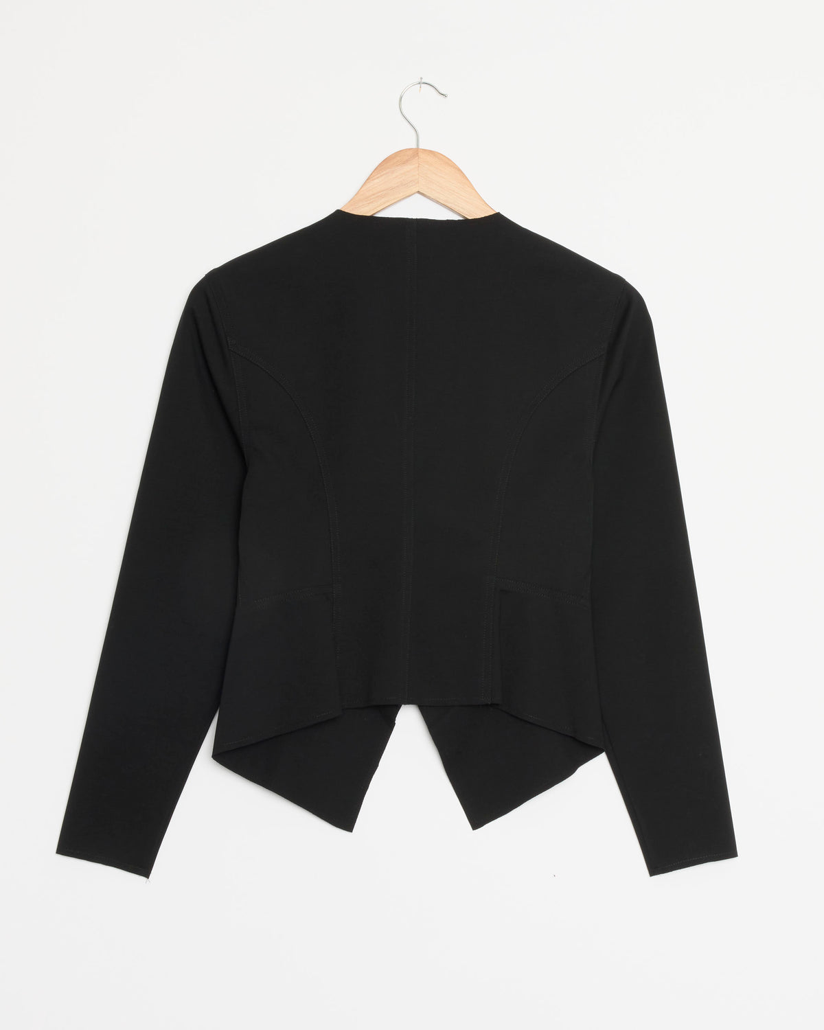 The Demi Jacket in Ink by Chelsea Mak is a black tailored jacket featuring long sleeves and a pointed hem at the back, shown on a wooden hanger against a white background. For questions about sizing or fit, our online chat is available to assist you.