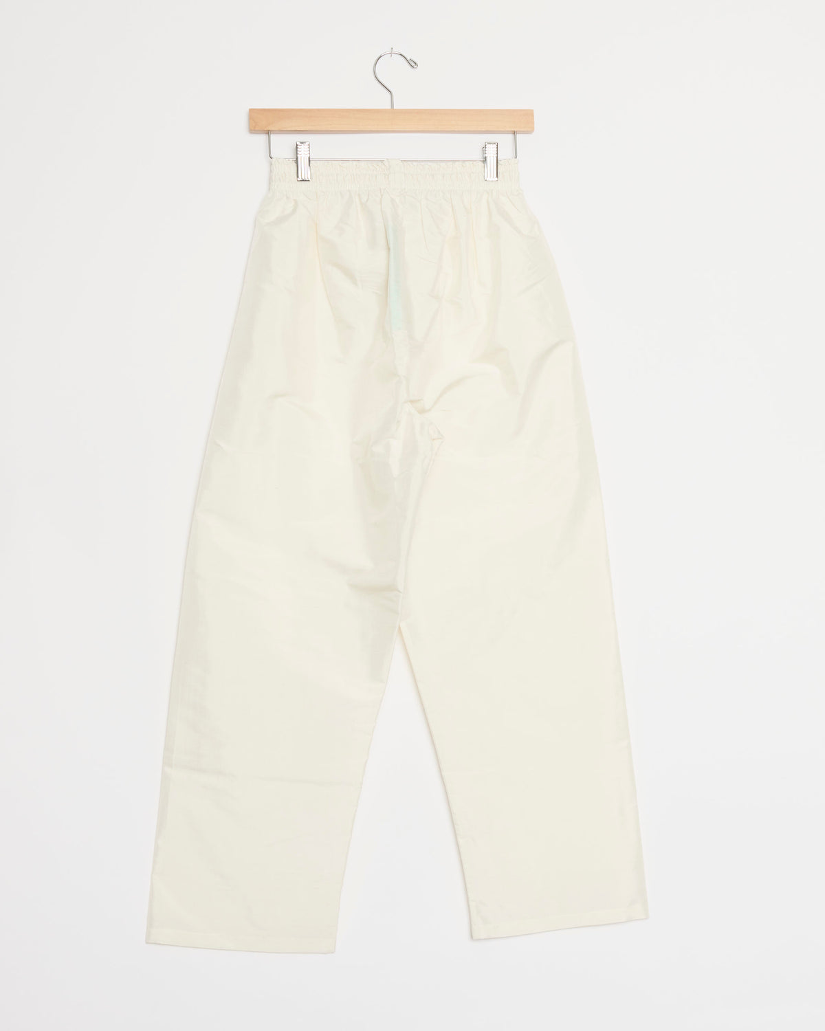 The Lexy Pant in White by Chelsea Mak, featuring a high-waisted design and elastic waistband, is displayed hanging on a wooden hanger against a plain white background.