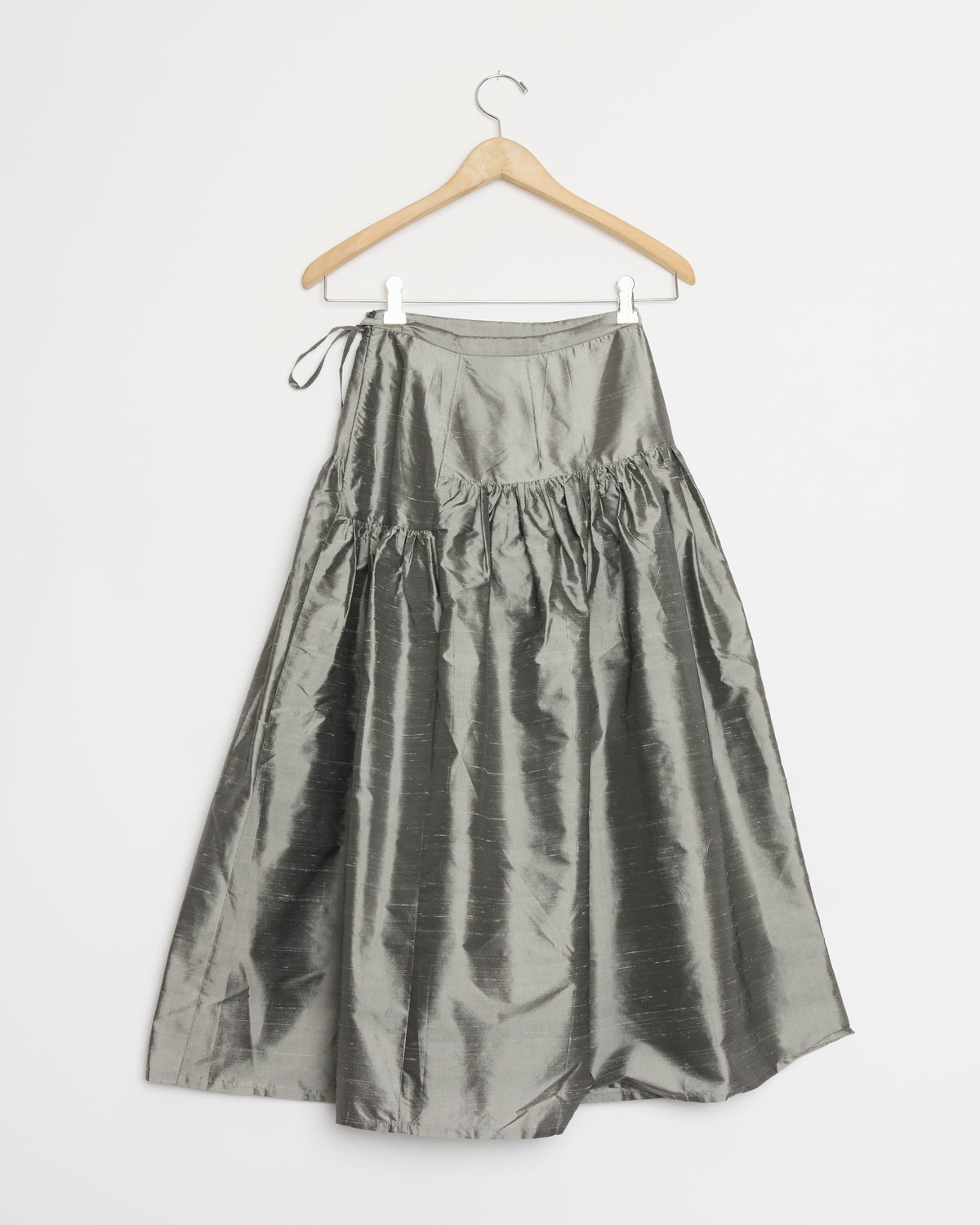 A Chelsea Mak Garden Skirt in Metal, featuring silver shiny silk taffeta with a gathered waist, is displayed on a wooden hanger against a plain white background.