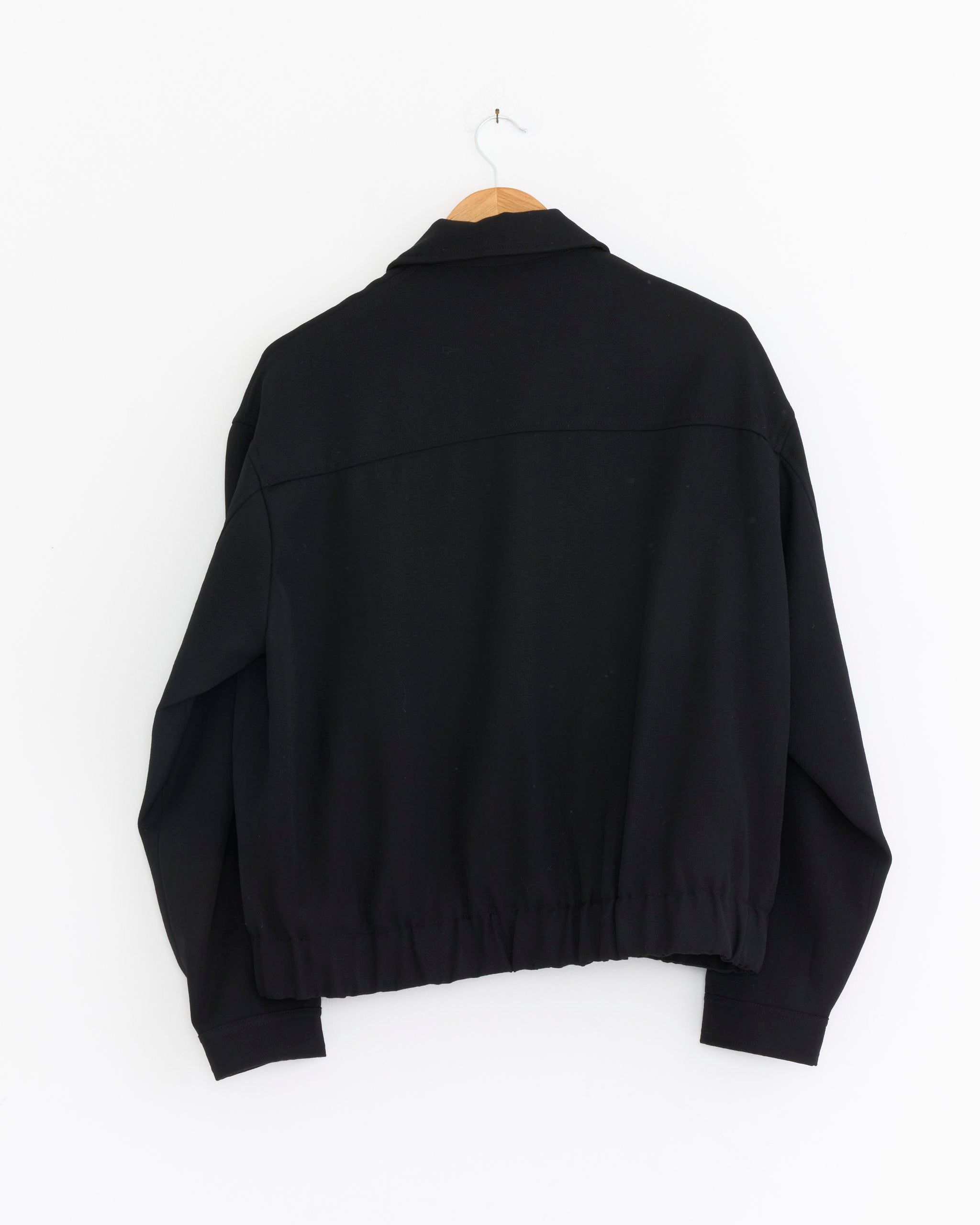 The James Coward Machine Jacket in Black, crafted from Japanese wool gabardine with long sleeves, is displayed from the back on a wooden hanger against a plain white wall.