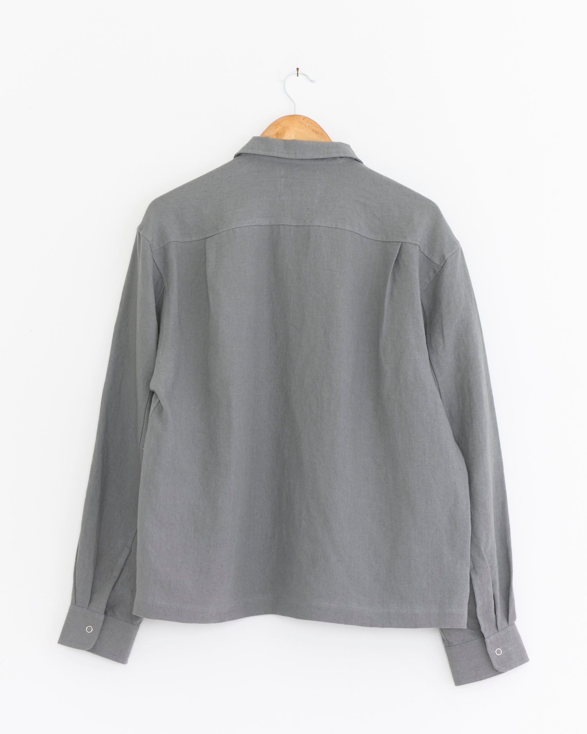 The James Coward Workshop Shirt in Flint, a gray Belgian linen long-sleeve work shirt, is displayed from the back on a wooden hanger against a plain white background.