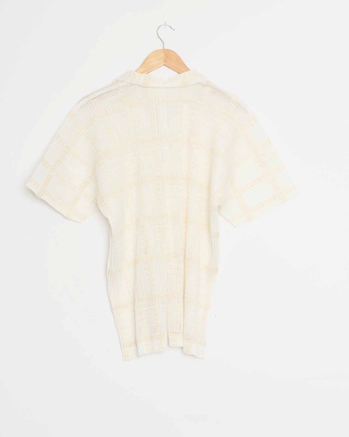 The Lace Grid Pleats Shirt in White by Homme Plissé Issey Miyake, featuring short sleeves and light grid pleats, is displayed from the back on a wooden hanger against a plain white background. The subtle pleated detailing highlights the brand’s signature style.
