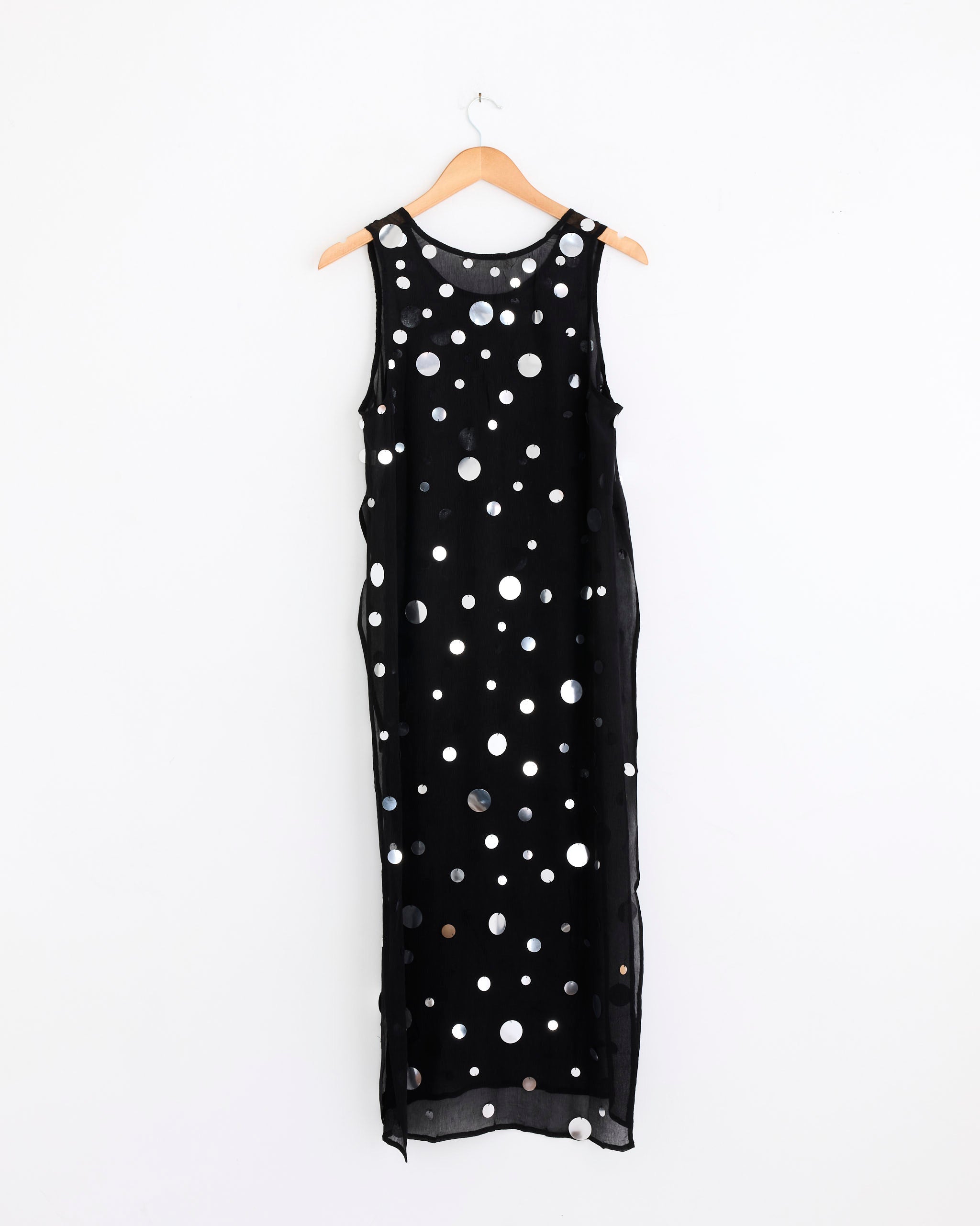 The Chloe Sequin Tunic in Black by Chan Luu, a sleeveless silk chiffon piece with a long straight cut and shiny silver polka dots, hangs on a wooden hanger against a plain white wall.