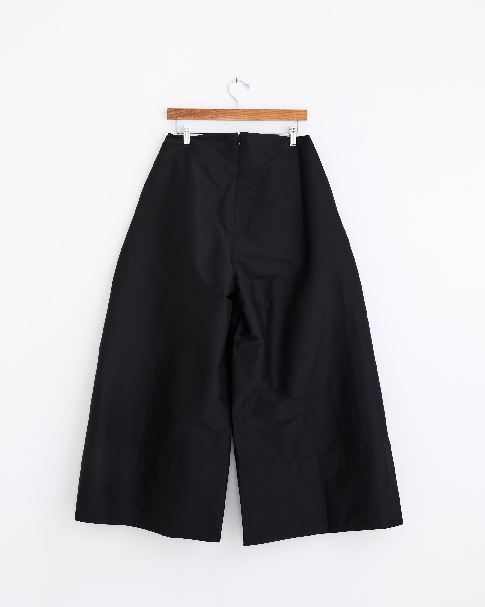The Chan Luu Techno Taffeta Ultra Wide Leg Trouser in Black hangs on a wooden hanger against a plain white wall.