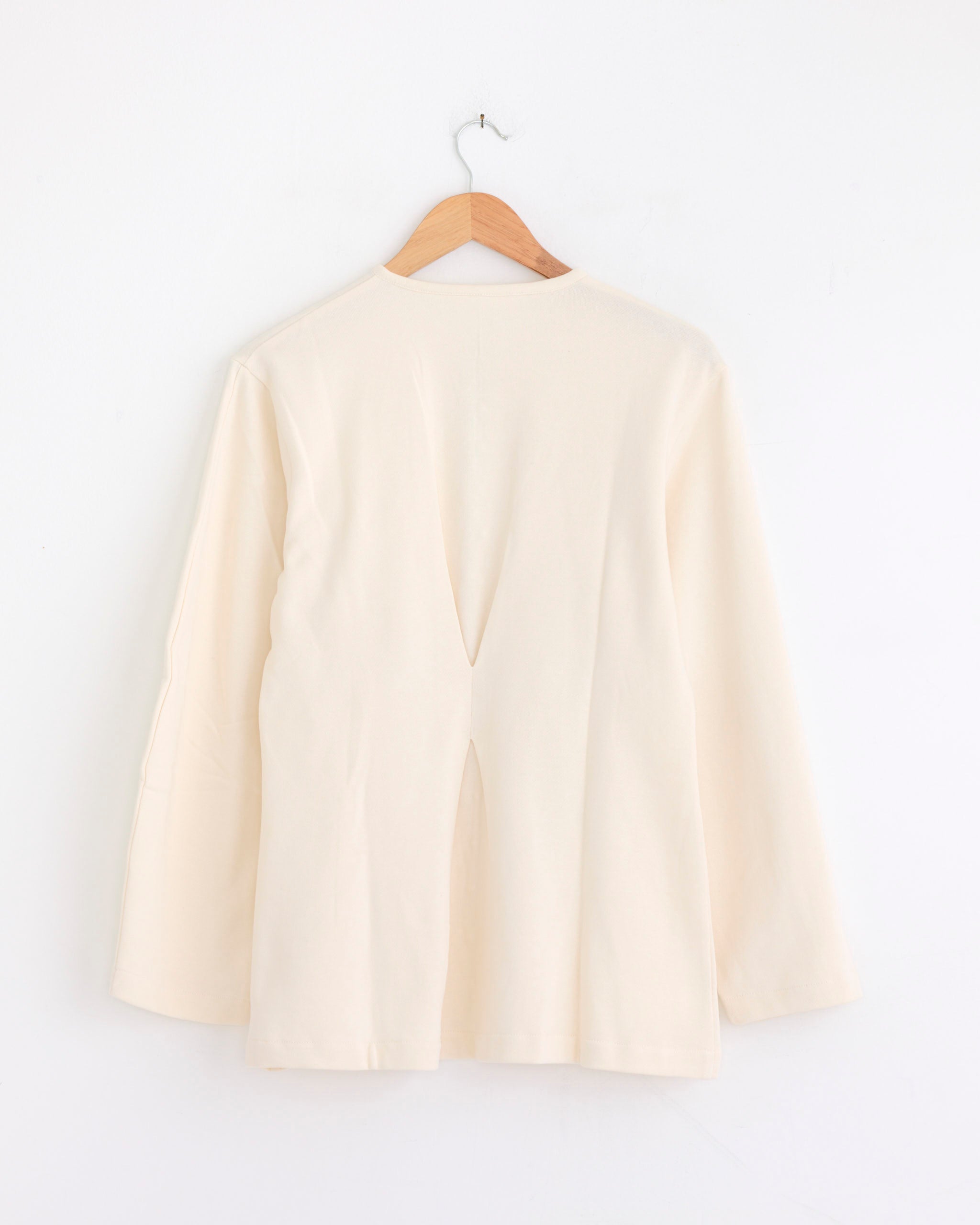 The Chan Luu Pinch Waist Cardigan in Ivory hangs on a wooden hanger against a white wall, displayed with the back facing forward. It features subtle balloon sleeves and a center slit detail on the back.