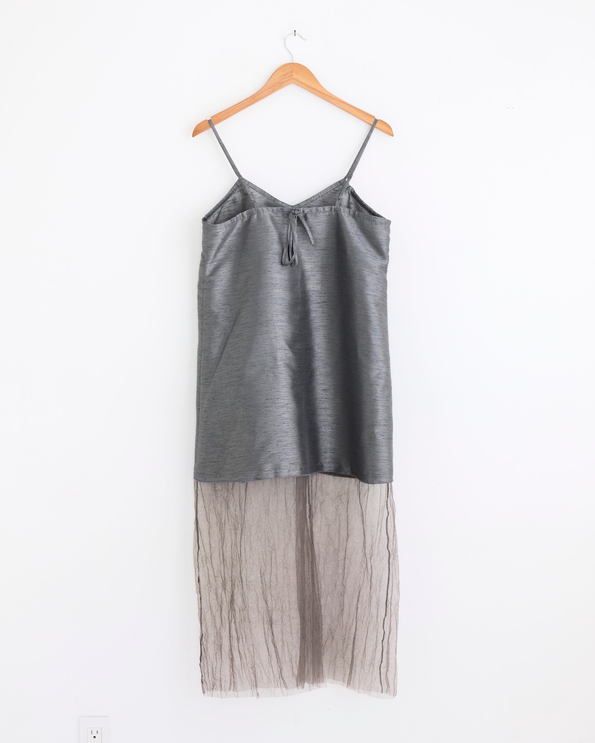 The Chan Luu Dupioni Mesh Dress in Steel, featuring thin straps, a satin-like top, and a sheer mesh skirt, hangs on a wooden hanger against a white wall.
