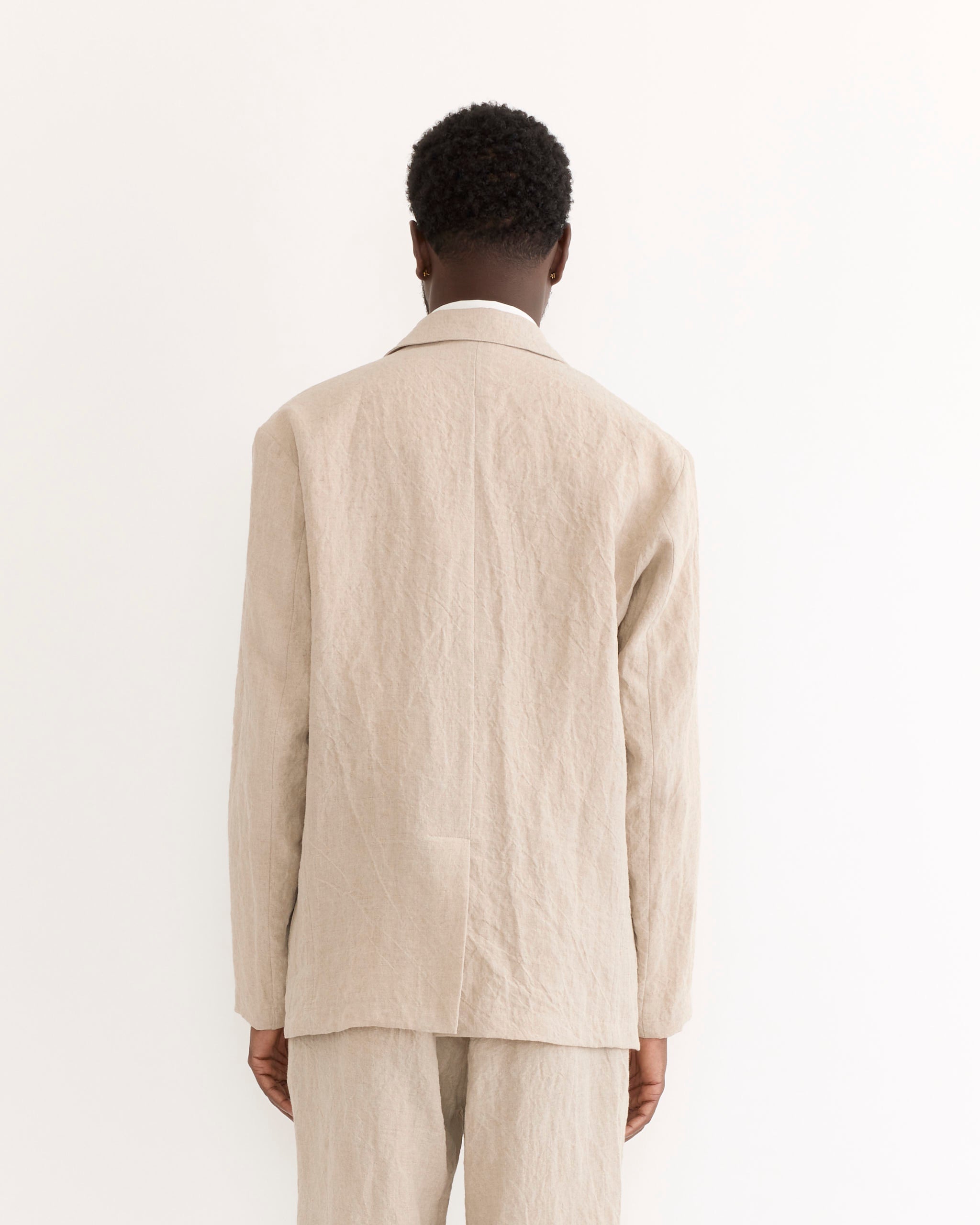 A person with short curly hair faces away from the camera, wearing the SMOCK Blazer in Sun Dried Linen Ivory against a plain white background.