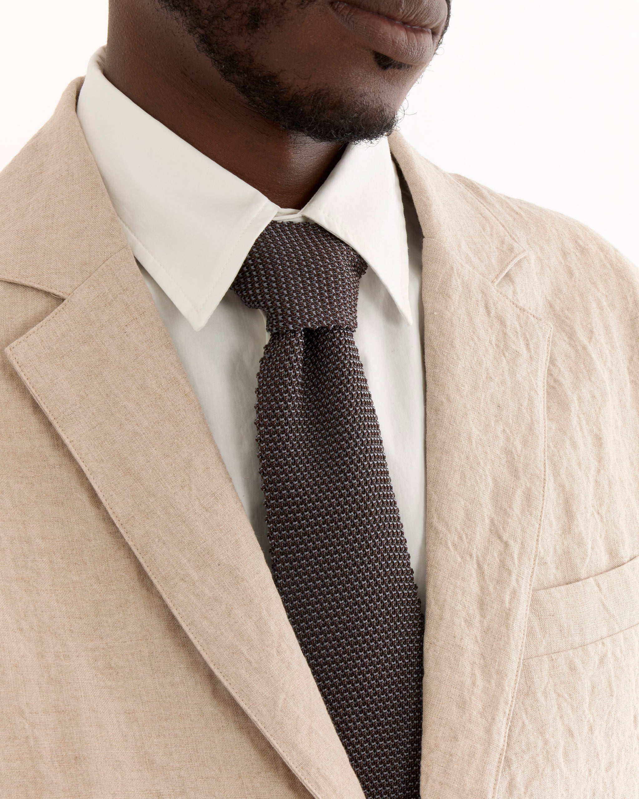 A close-up of a person wearing the SMOCK Blazer in Sun Dried Linen Ivory, paired with a white dress shirt and dark textured knit tie. The image highlights the upper torso, with the person's eyes out of frame.