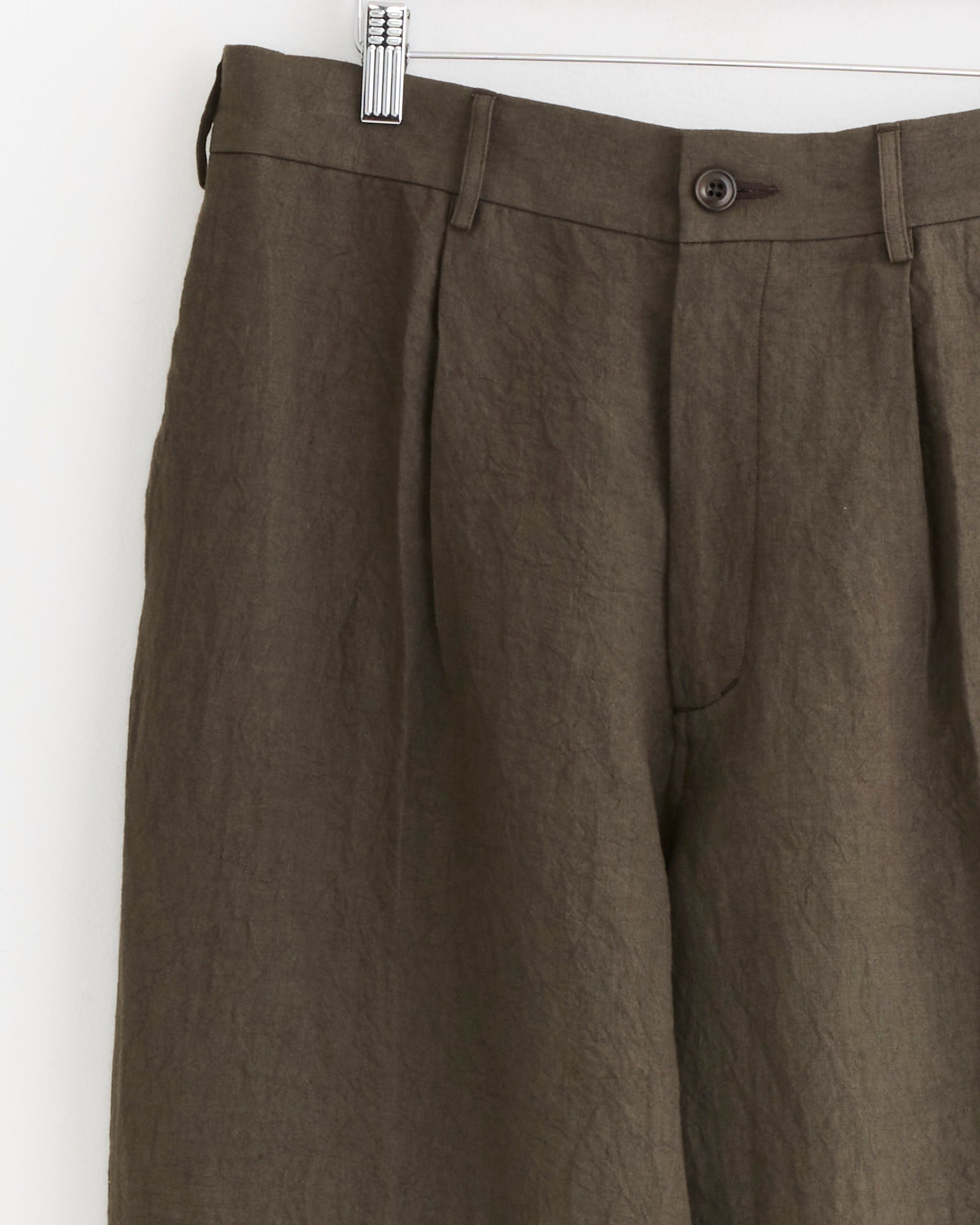 A pair of SMOCK West Coast Pant in Sun Dried Linen Tobacco, featuring Japanese linen, button and zip closure, pleated front, and belt loops, hangs on a metal hanger against a white background.
