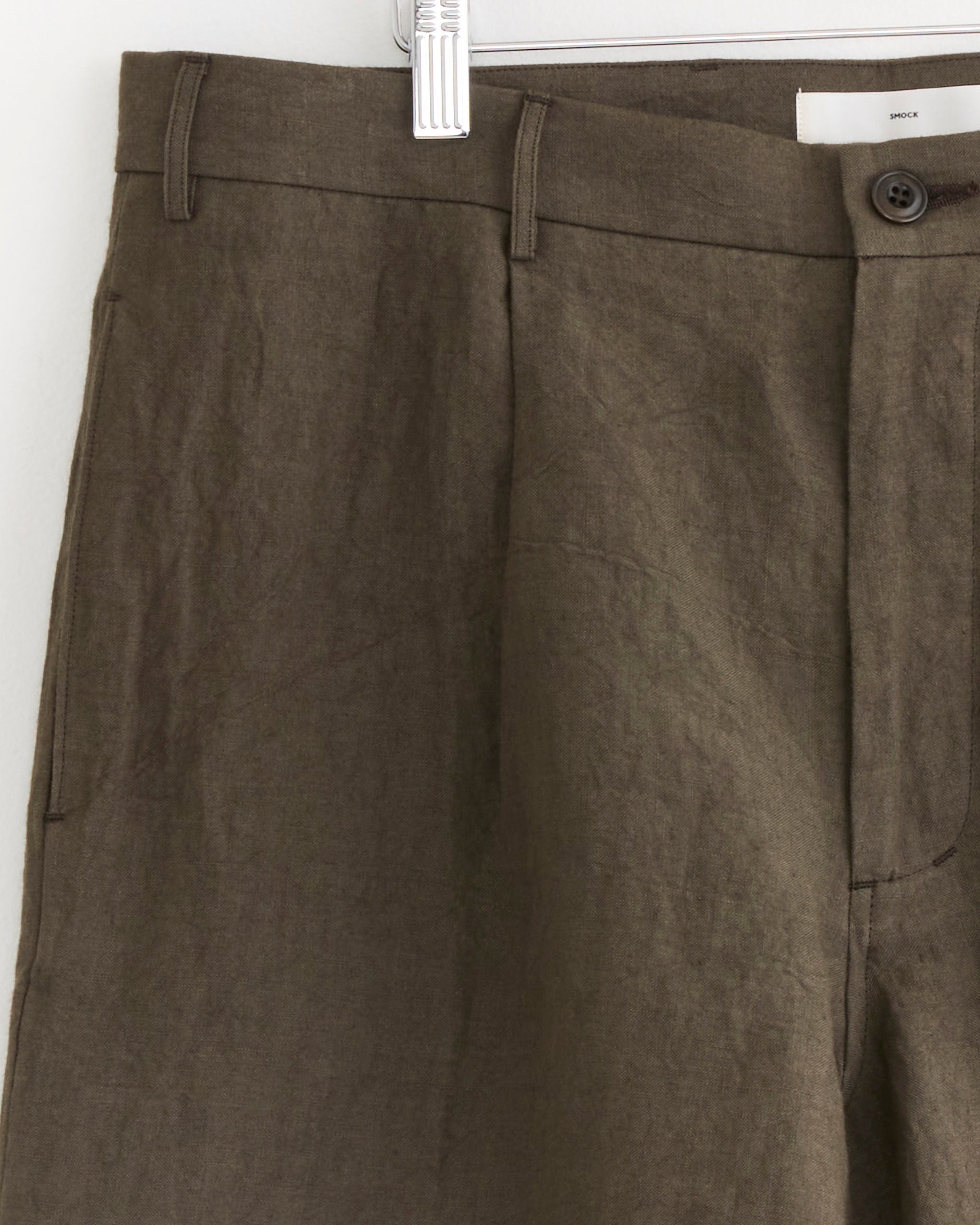 A close-up of the Arroyo Short in Sun Dried Linen Tobacco by SMOCK, hanging on a hanger, highlights the waistband, button, belt loop, and front pockets.