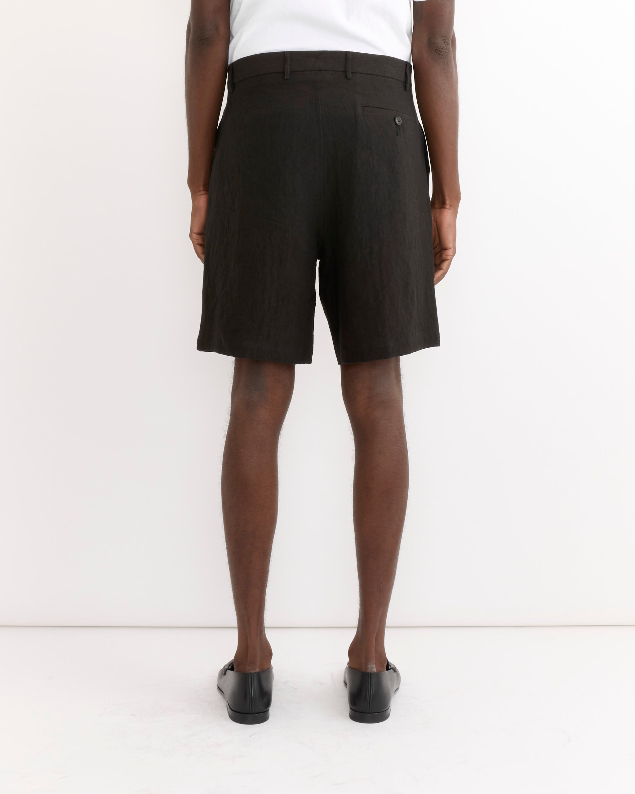 A person faces away from the camera against a white background, wearing a white top and SMOCK Arroyo Short in Sun Dried Linen Black with black slip-on shoes.