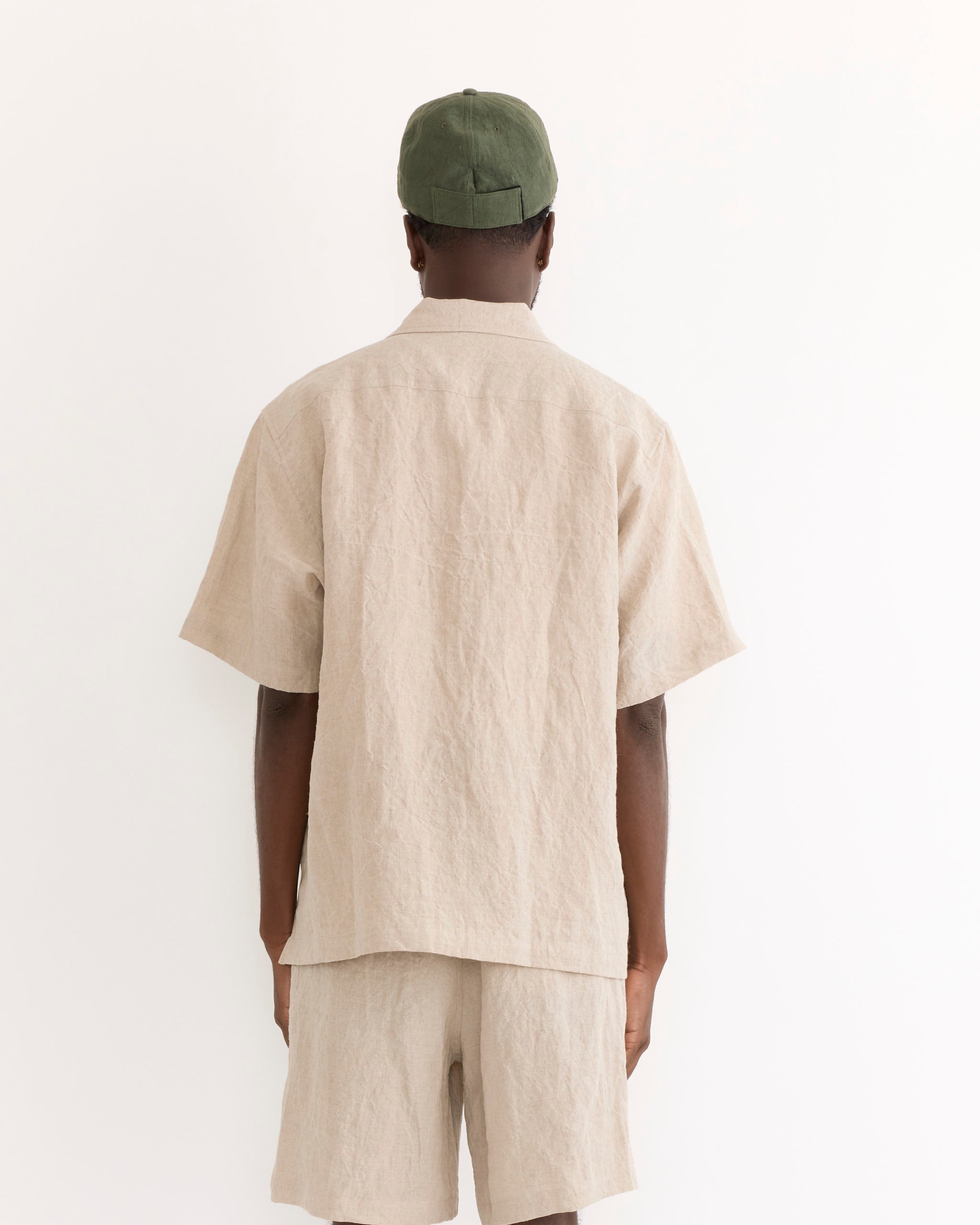 A person stands facing away, wearing the SMOCK Cardiff Shirt in Sun Dried Linen Ivory with matching shorts and a green cap, set against a plain white background.