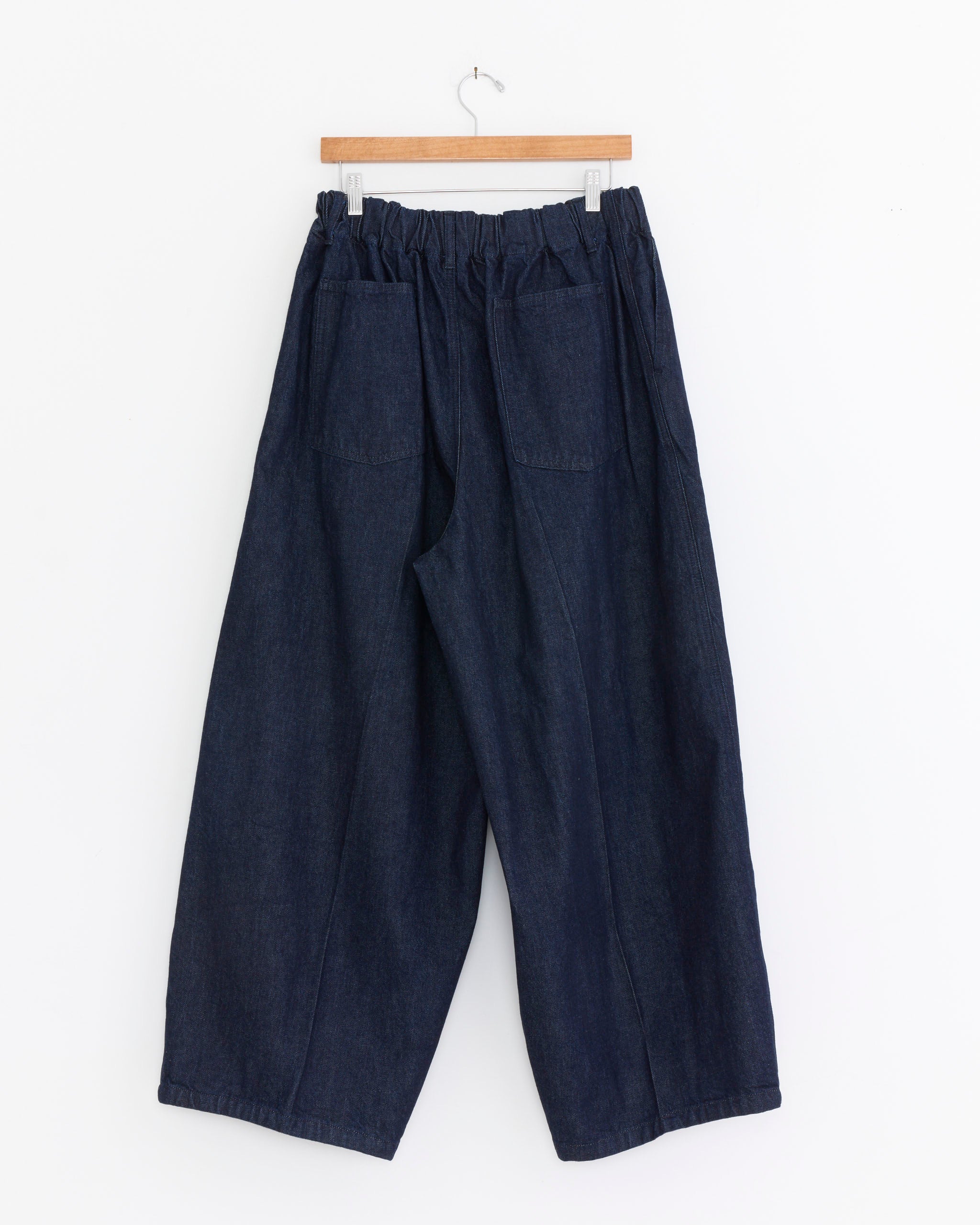 The Sillage Circular Denim Pant in One Wash, crafted from dark blue denim with an elastic waistband and two large front pockets, hangs on a wooden hanger against a white background. Made in Japan.