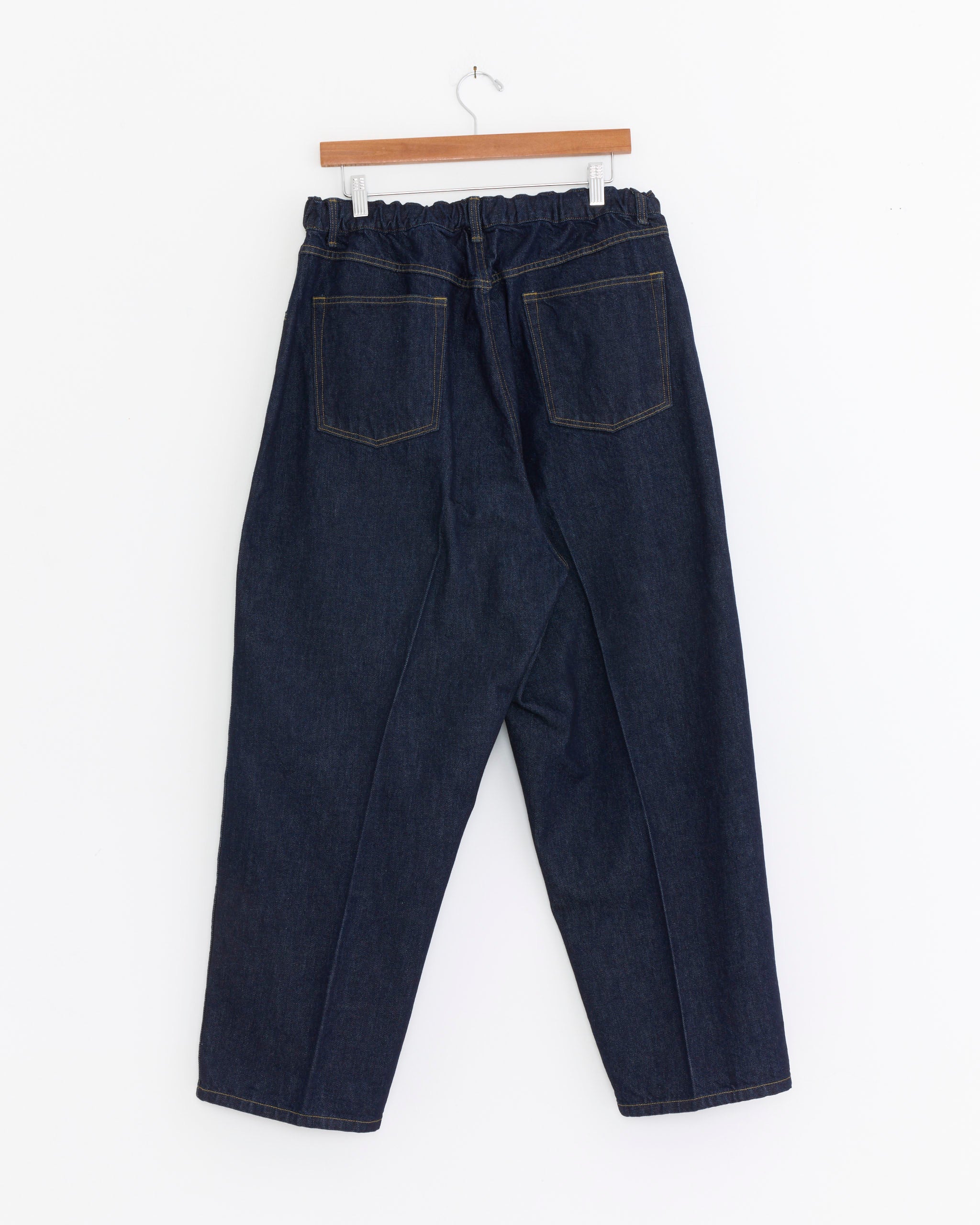 The Sillage Essentials 5 Pocket Denim Pant in One Wash features a dark blue relaxed tapered silhouette, organic cotton fabric, elastic waistband, and two back pockets. Made in Japan and shown on a wooden hanger against a white background.