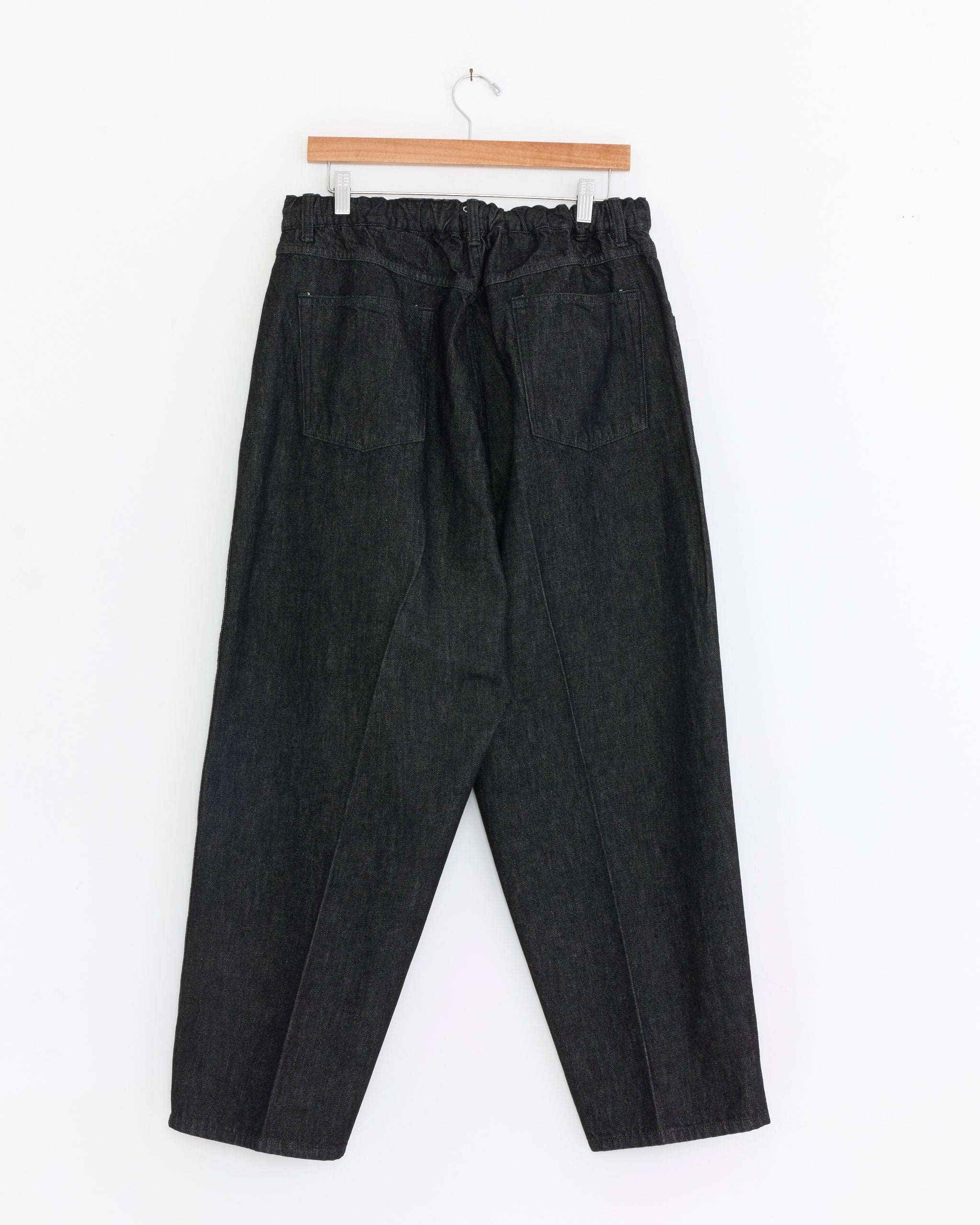 The Sillage Essentials 5 Pocket Denim Pant in Black One Wash is a high-waisted, loose-fit jean with an elastic waistband, made from organic cotton denim and displayed on a wooden hanger against a white background.