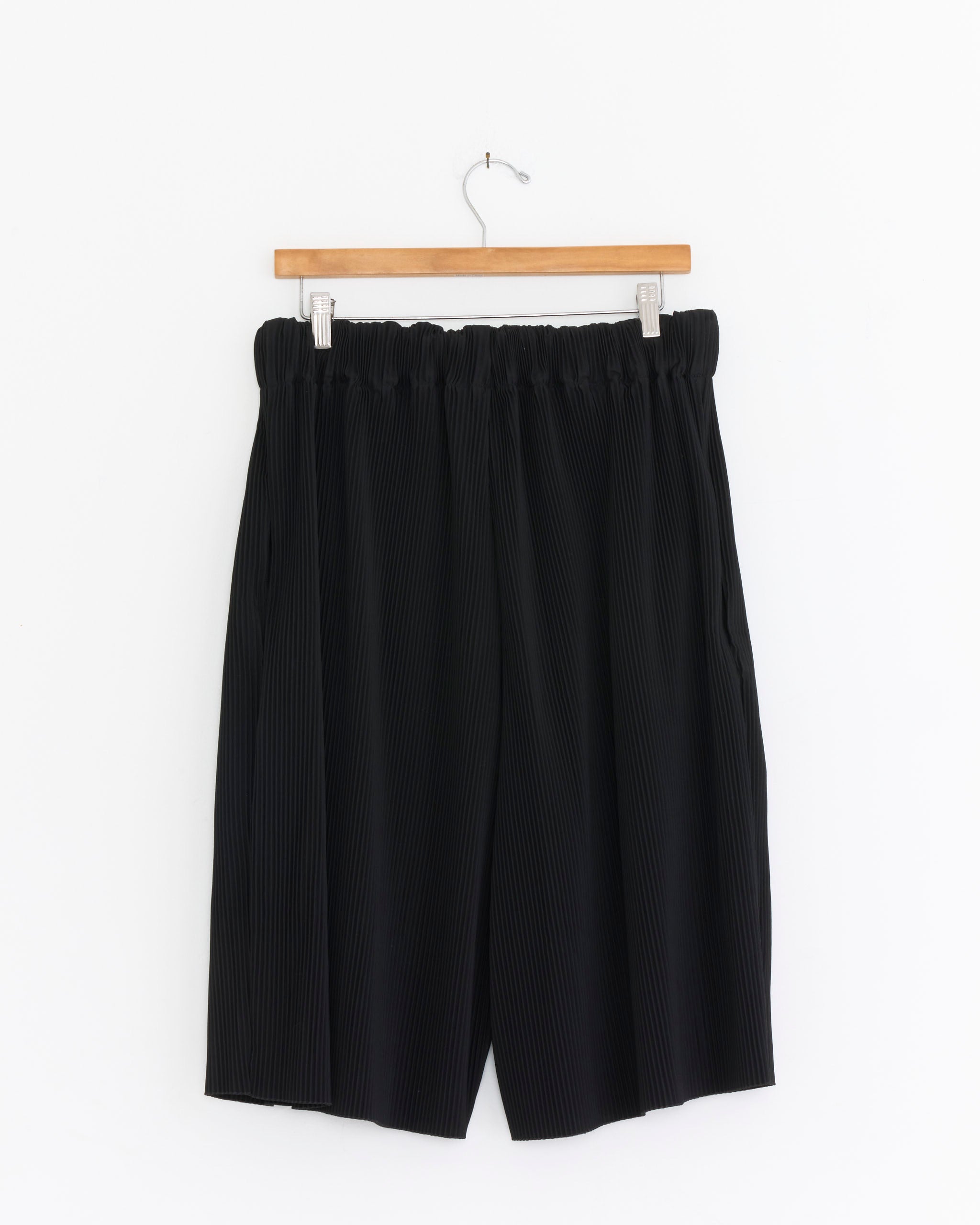 The Sillage Circular Short Pockets Pleat Pant in Black, featuring an elastic waistband and pleated design, is displayed on a wooden hanger against a plain white background.